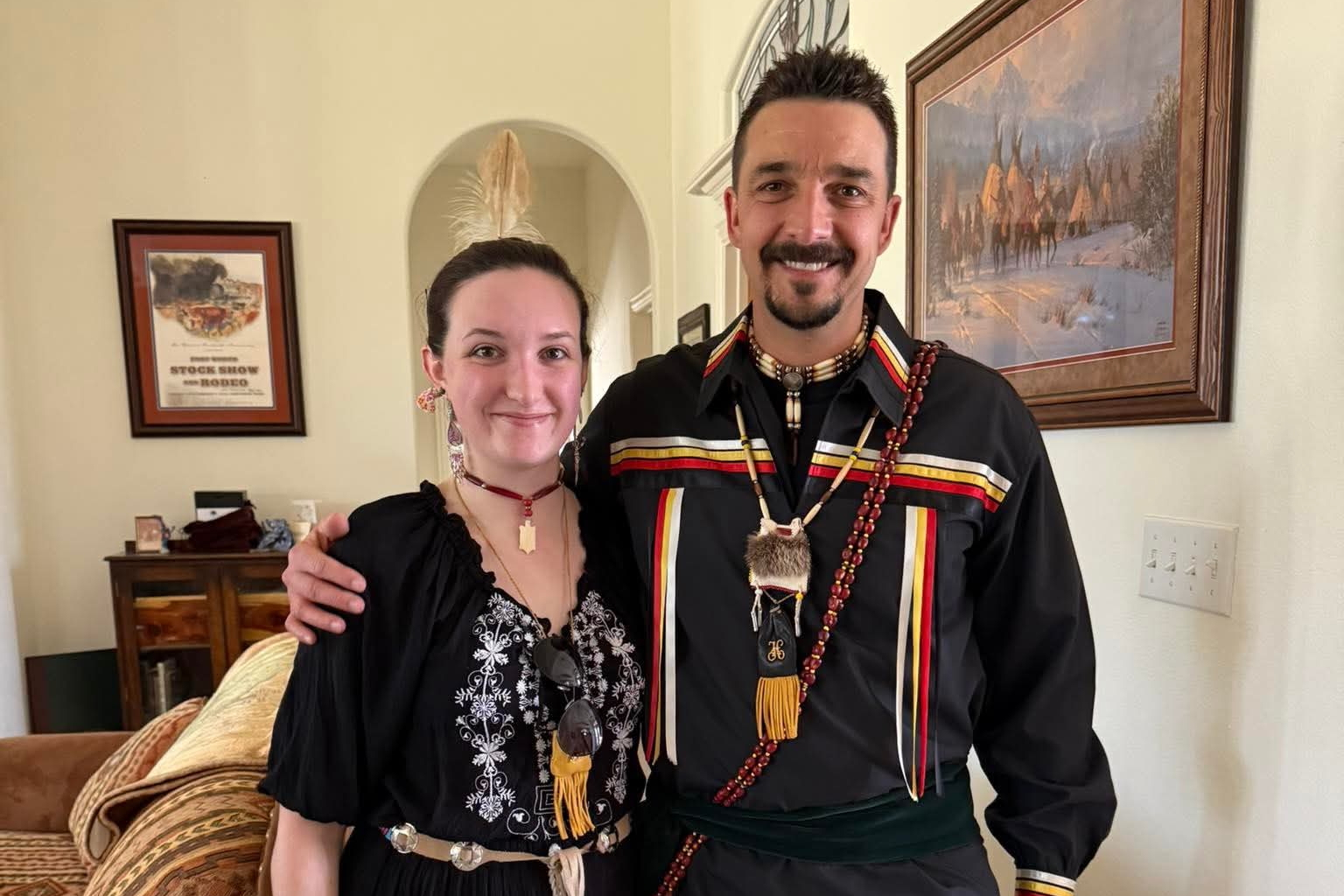 Two people in traditional-style clothing pose together in a living room, smiling at the camera.