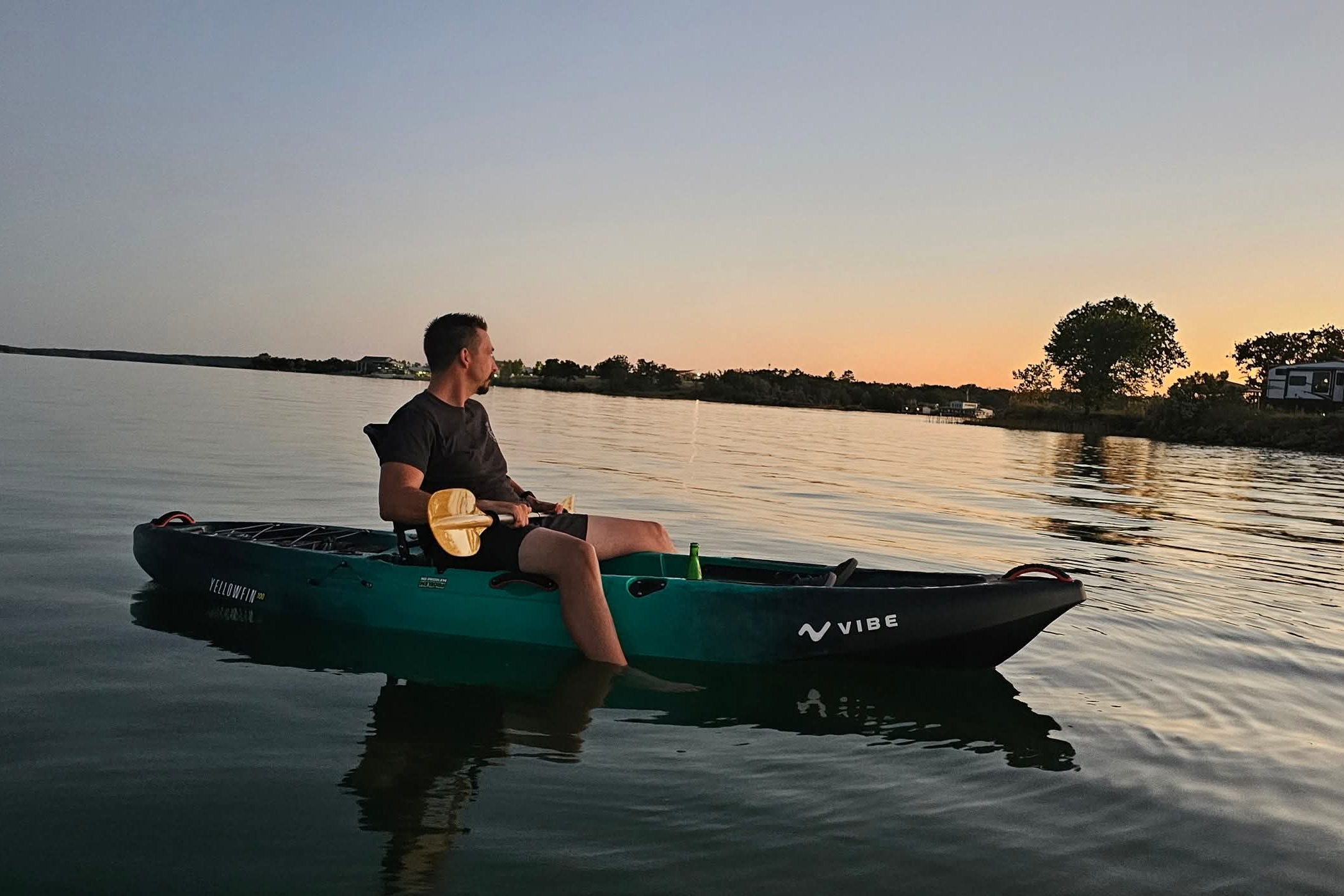Man in a green kayak on a calm lake at sunset, holding a wooden paddle across his lap with trees and houses on the horizon.
