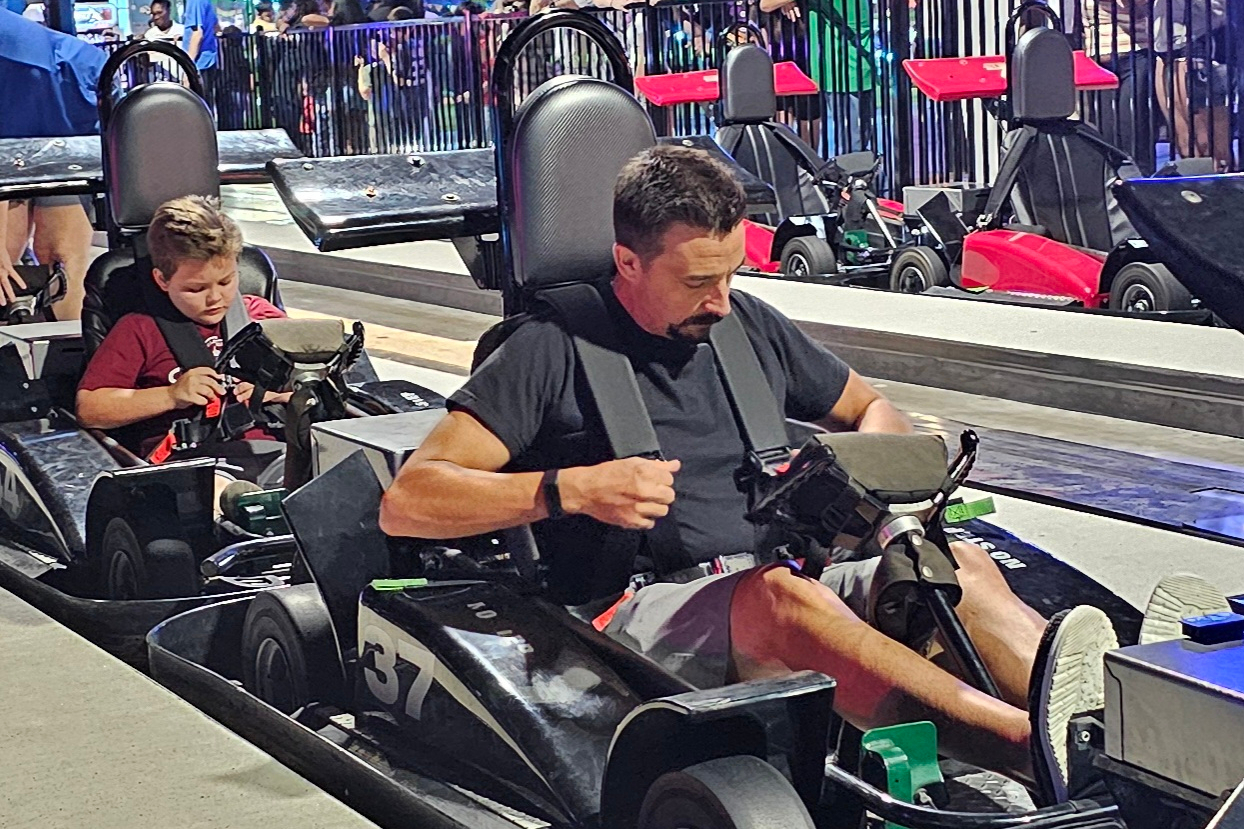 Man and child seated in go-karts at an indoor racing track, foreground kart numbered 57, ready to race.