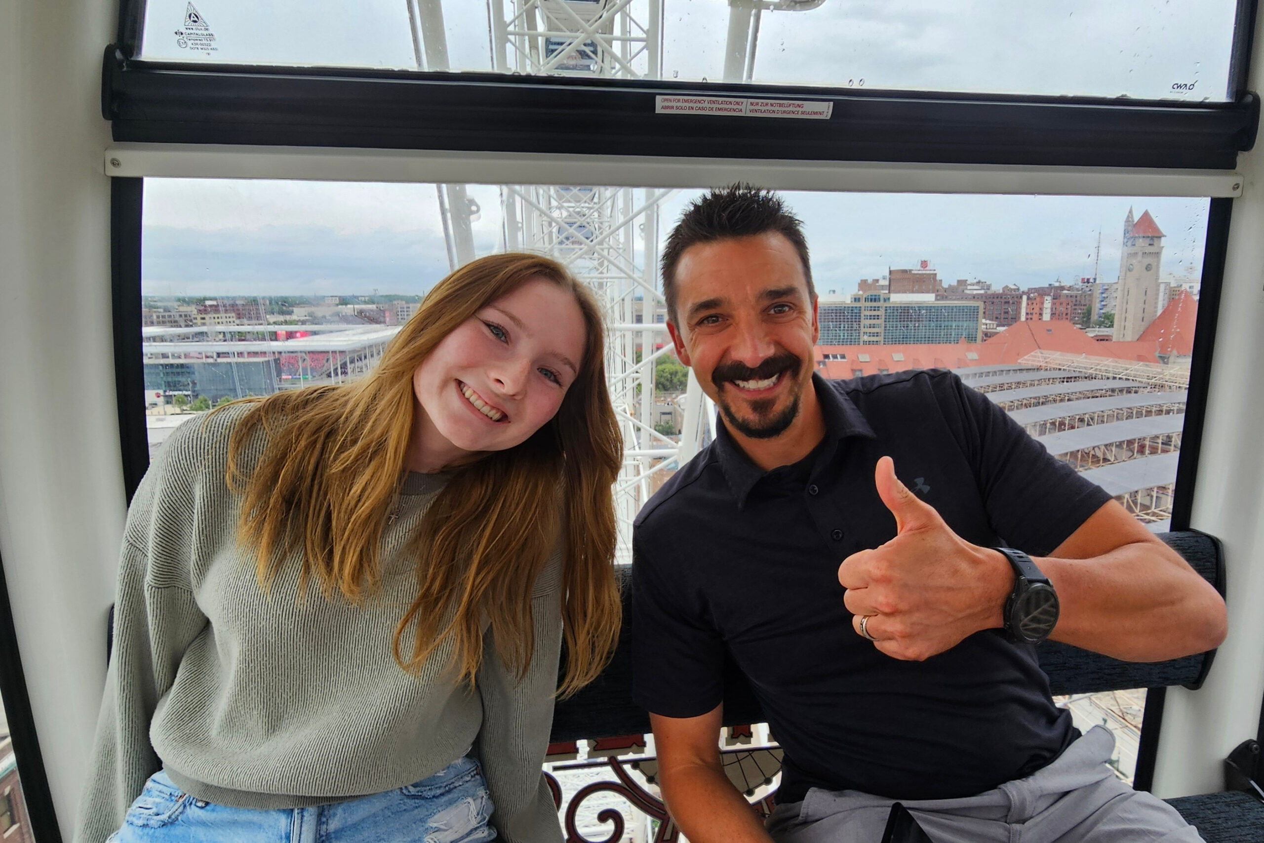 Smiling couple posing for a selfie inside a Ferris wheel cabin, with a city skyline and red-roofed buildings in the background.