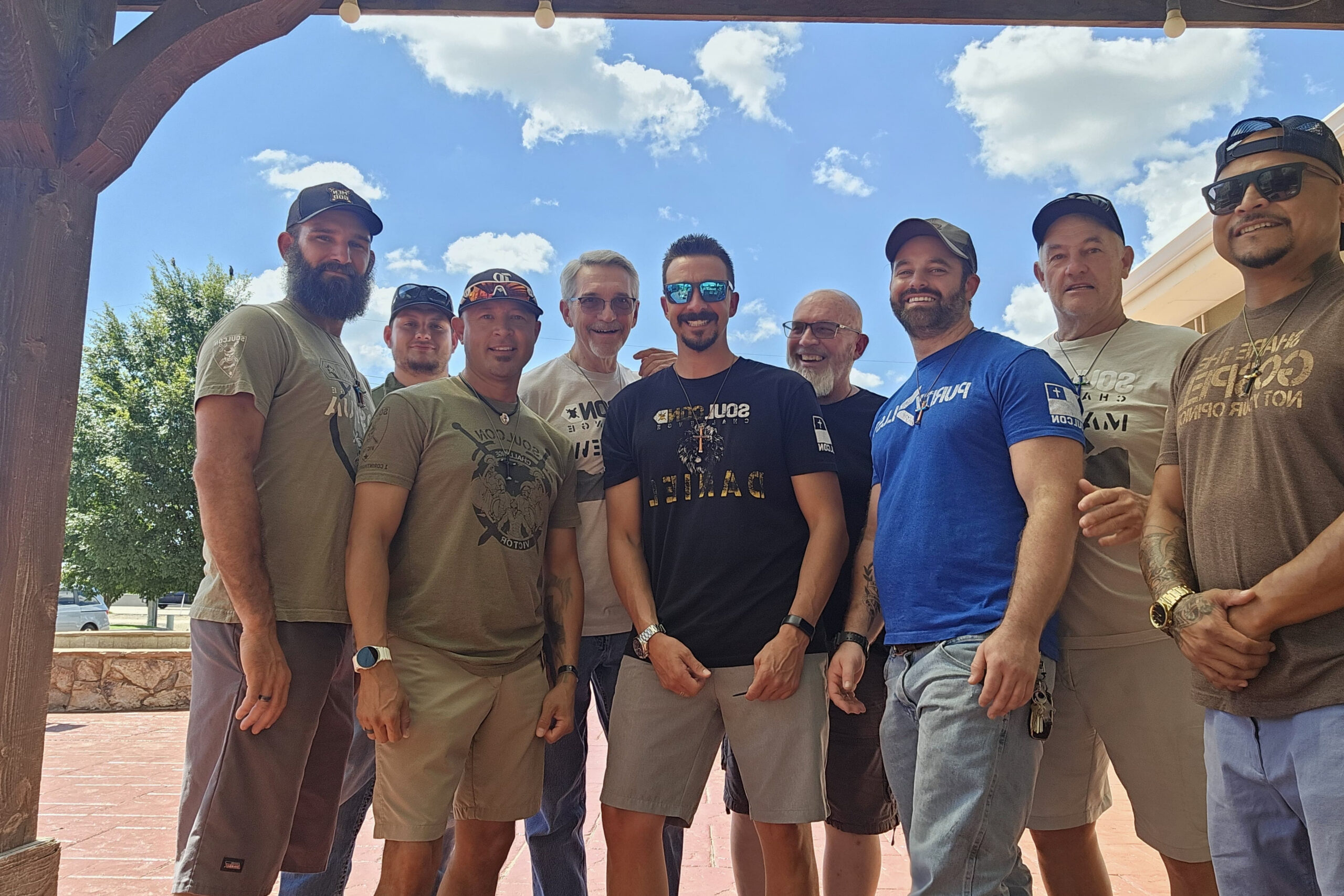 Group of men standing together outdoors under a wooden pergola, smiling at the camera, wearing casual t-shirts and hats.