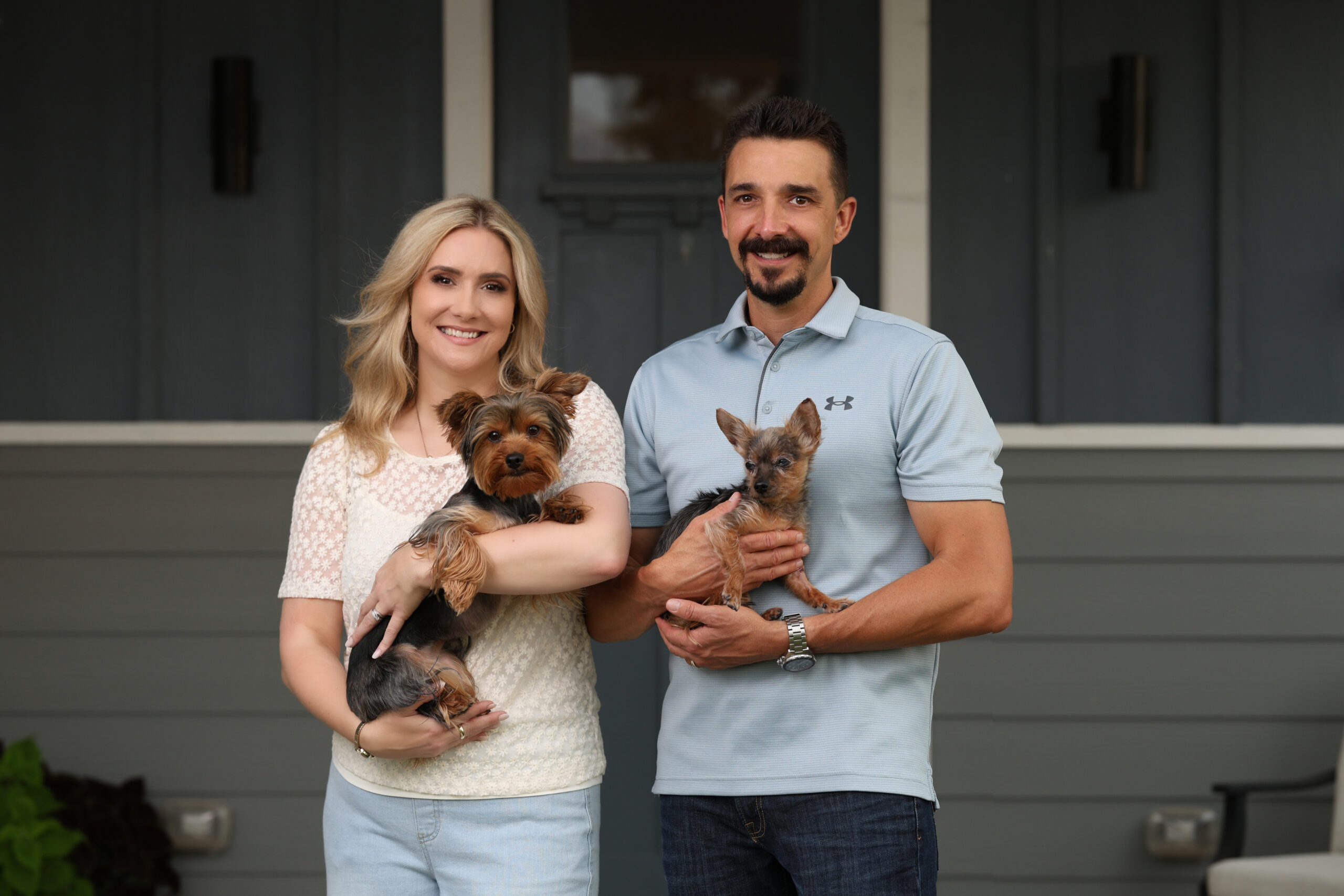 Smiling couple stands on a porch, each holding a small dog in front of a house.