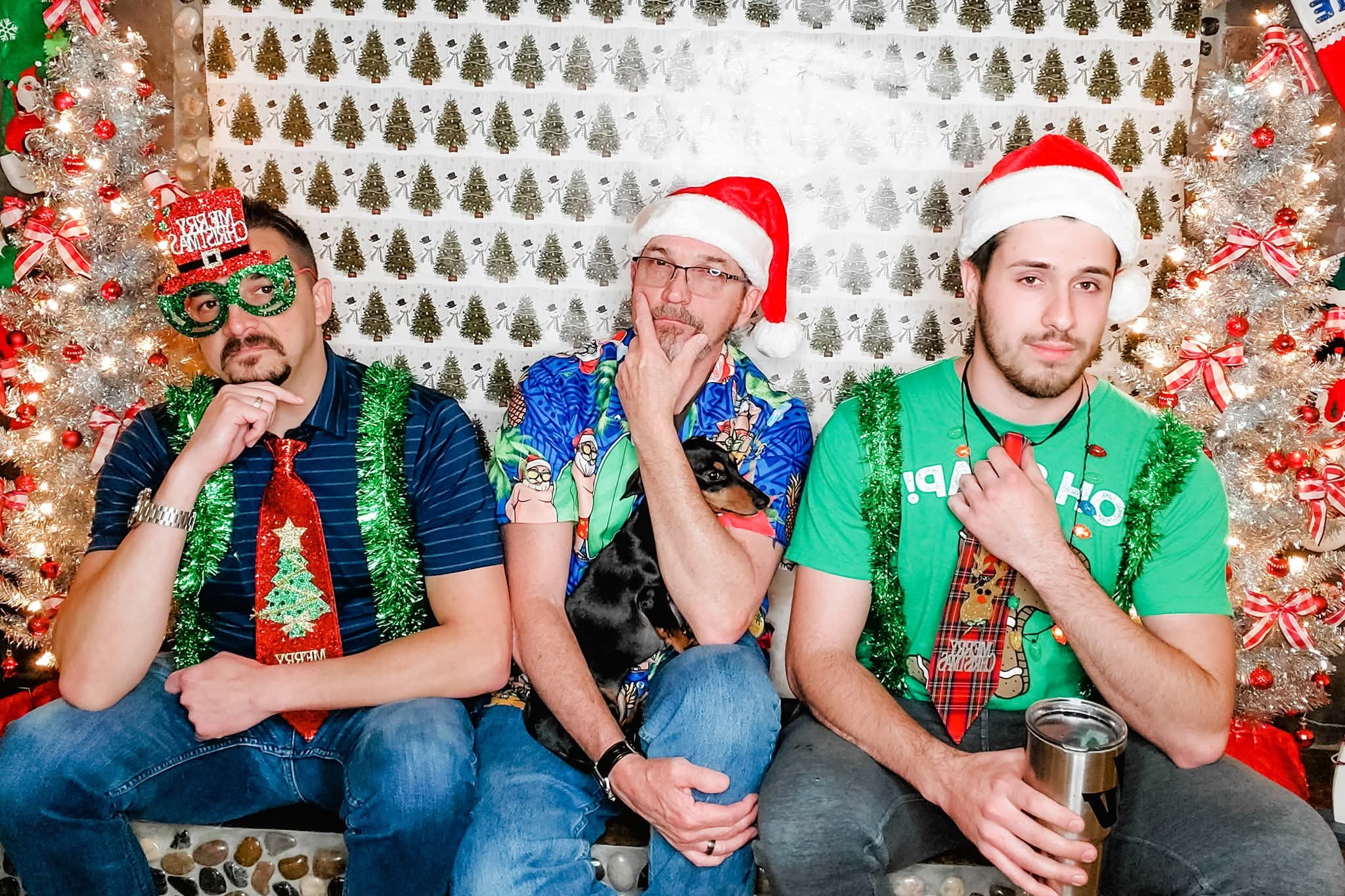 Three friends in Christmas outfits pose in a festive photo booth, wearing Santa hats, tinsel, and party accessories against a tree-pattern backdrop.