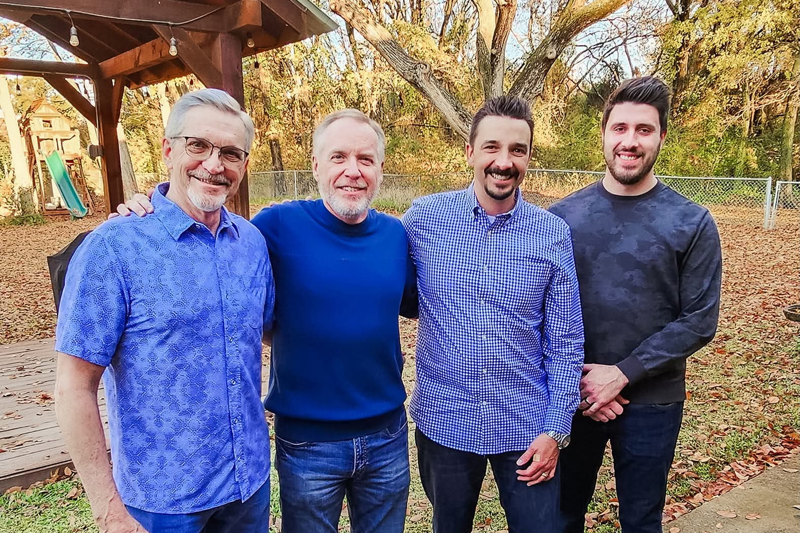 Four men posing together outdoors in a backyard, smiling, with autumn leaves on the ground and a wooden playset in the background.