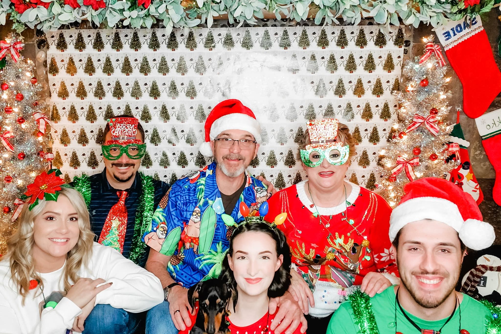 Group of people in colorful Christmas costumes and masks posing for a photo with a festive Christmas backdrop and lights behind them.