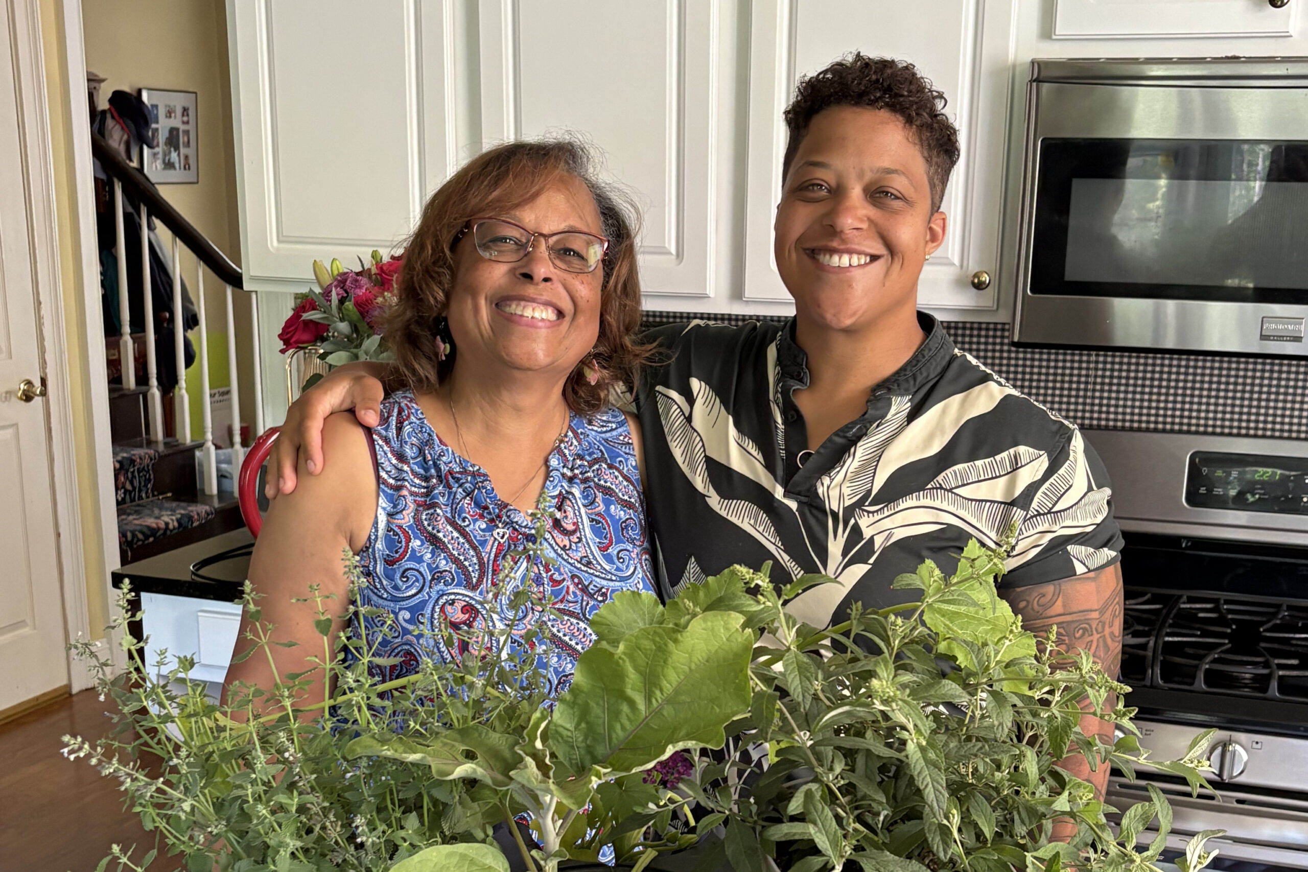 Two smiling women pose together in a bright kitchen, with leafy herbs in the foreground.