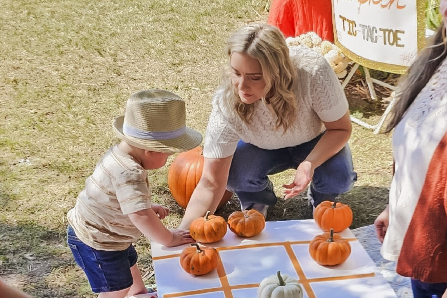 Woman helps a toddler place pumpkins on a tic-tac-toe board at an outdoor fall event.