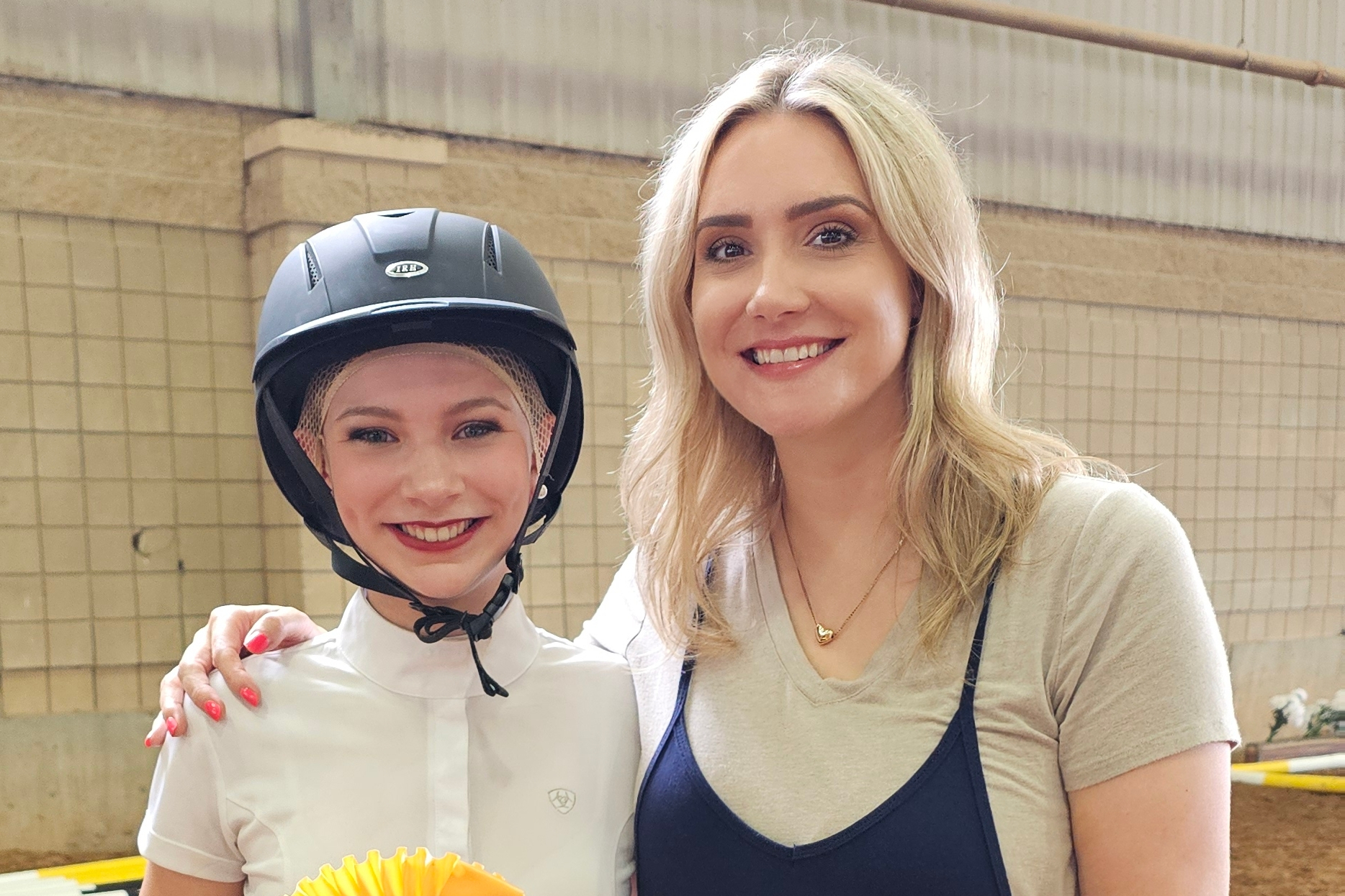 Smiling woman and young girl pose together indoors, the girl wearing a gray riding helmet and white shirt.