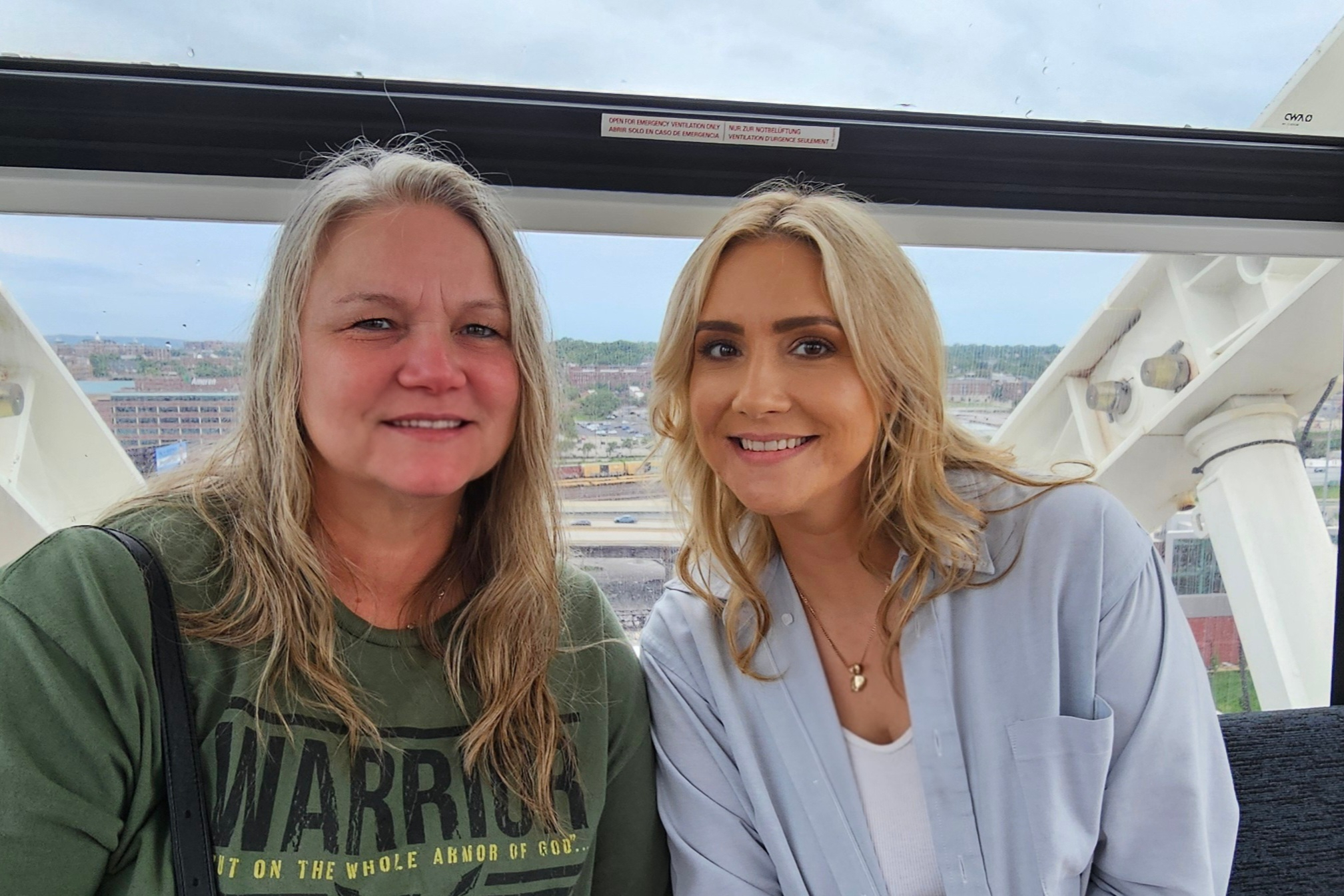 Two smiling women posing for a selfie inside a ferris wheel gondola with a city skyline in the background.