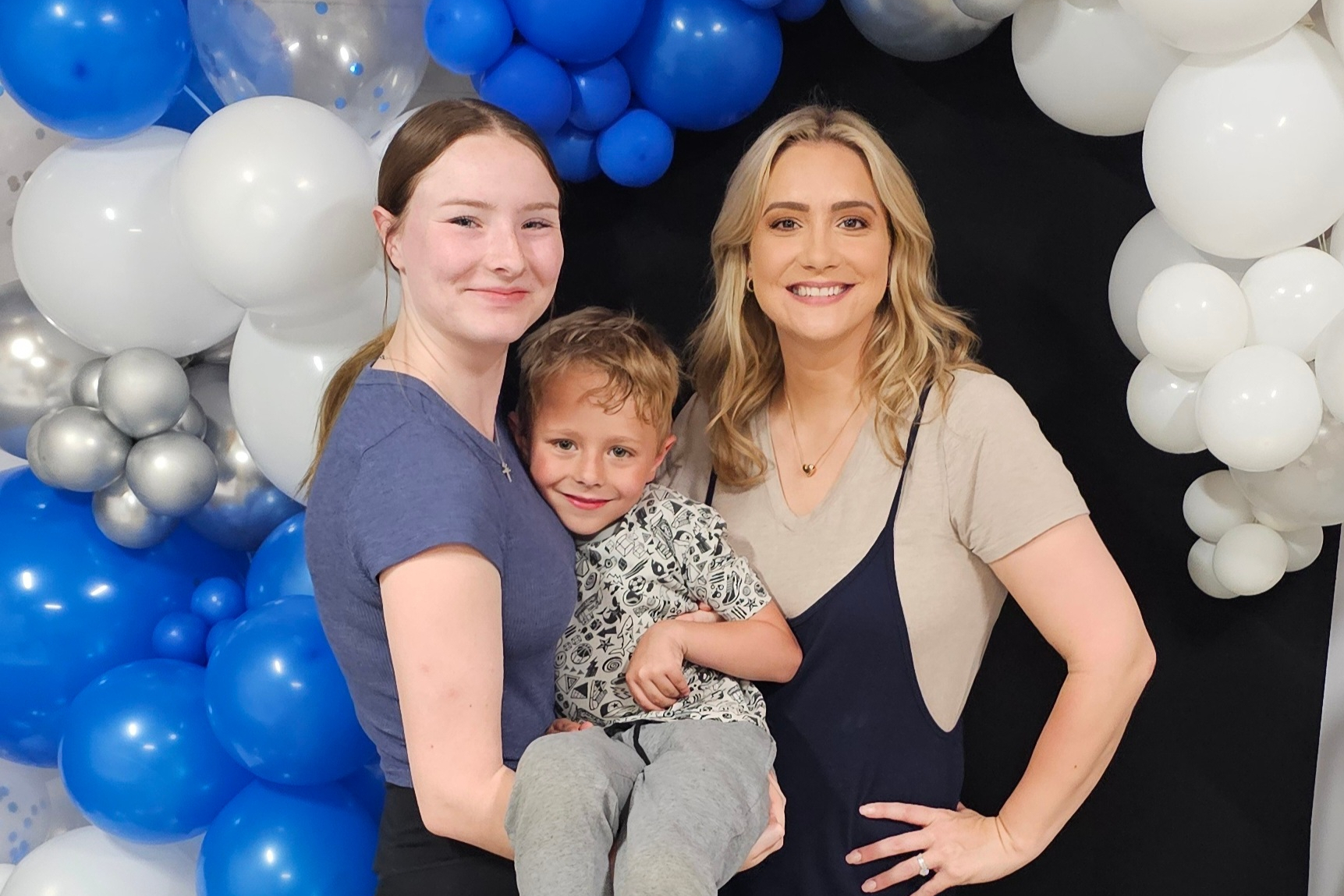 Three people smiling in front of a blue and white balloon wall backdrop (two women and a child).