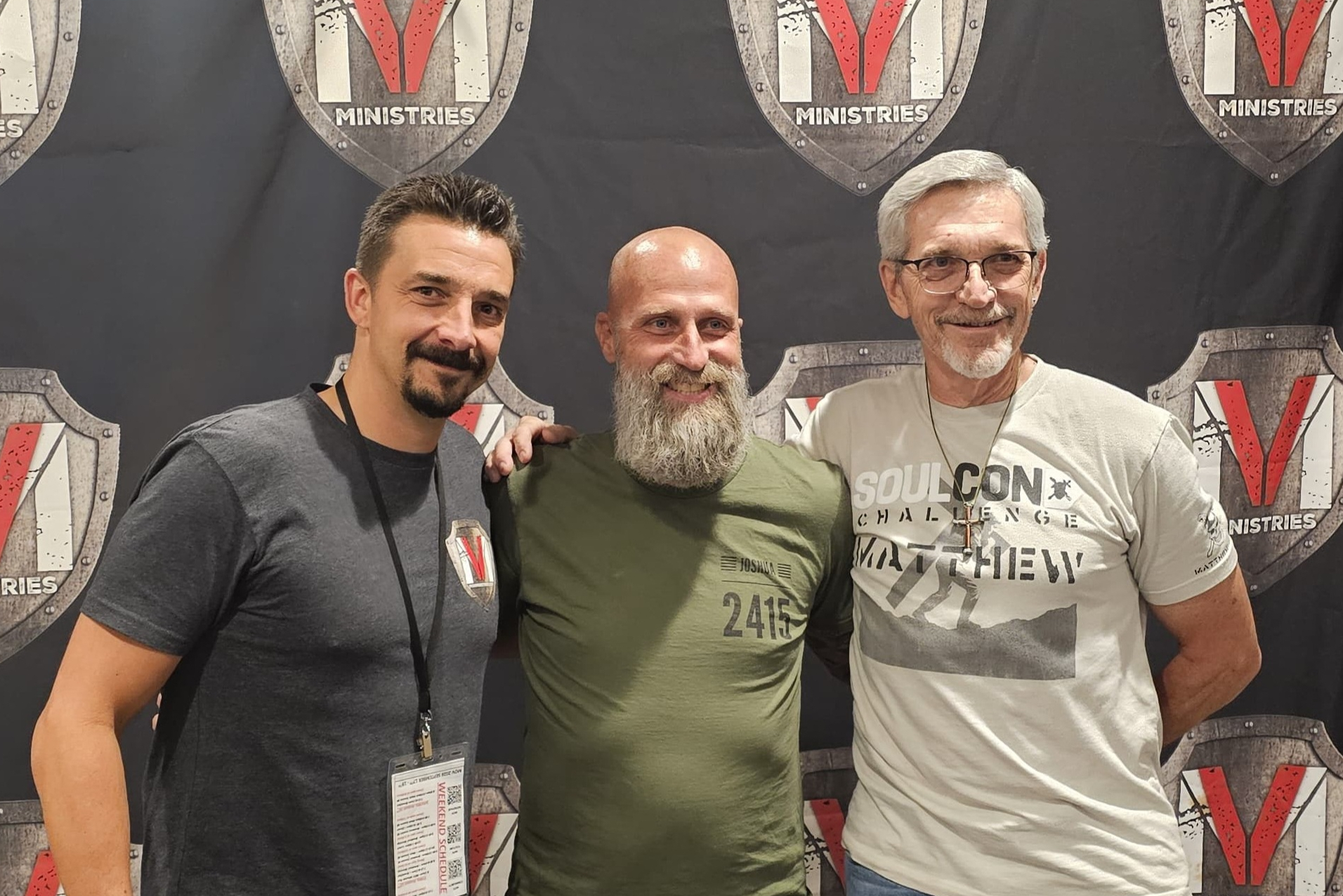 Three men posing together at a V Ministries event, smiling at the camera with badges visible on lanyards.