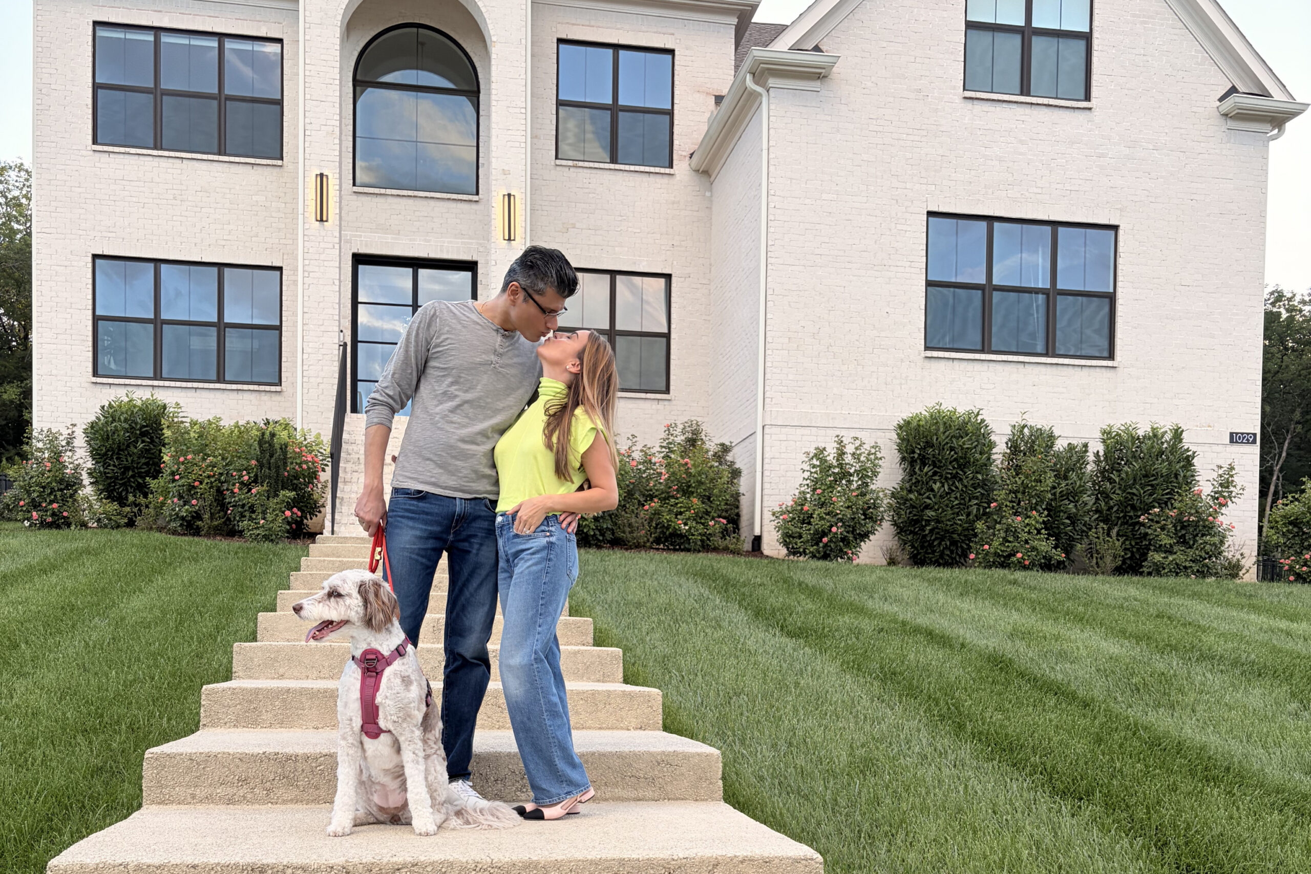 Couple kissing on the front steps of a beige brick house, with a white dog on a pink harness sitting at the bottom and a green lawn nearby.