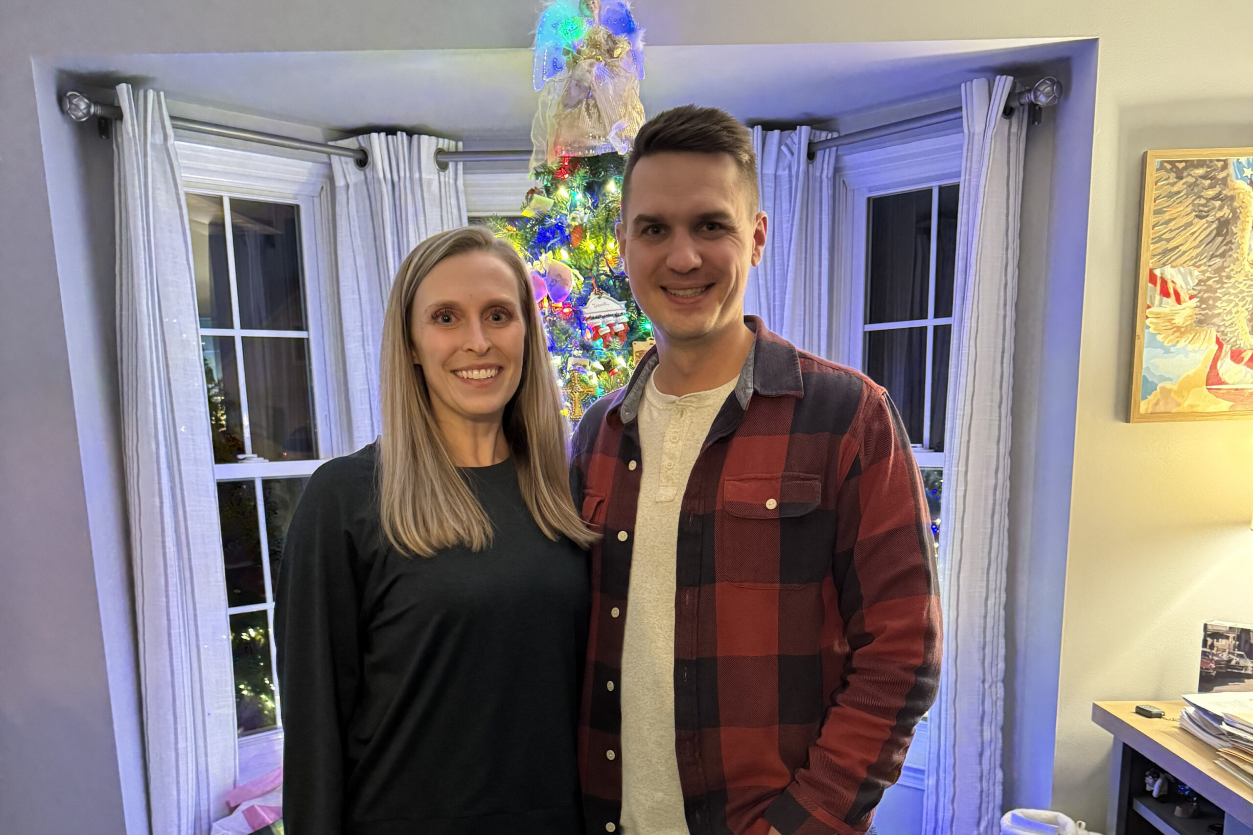 Smiling couple standing in a living room with a lit Christmas tree in the background.