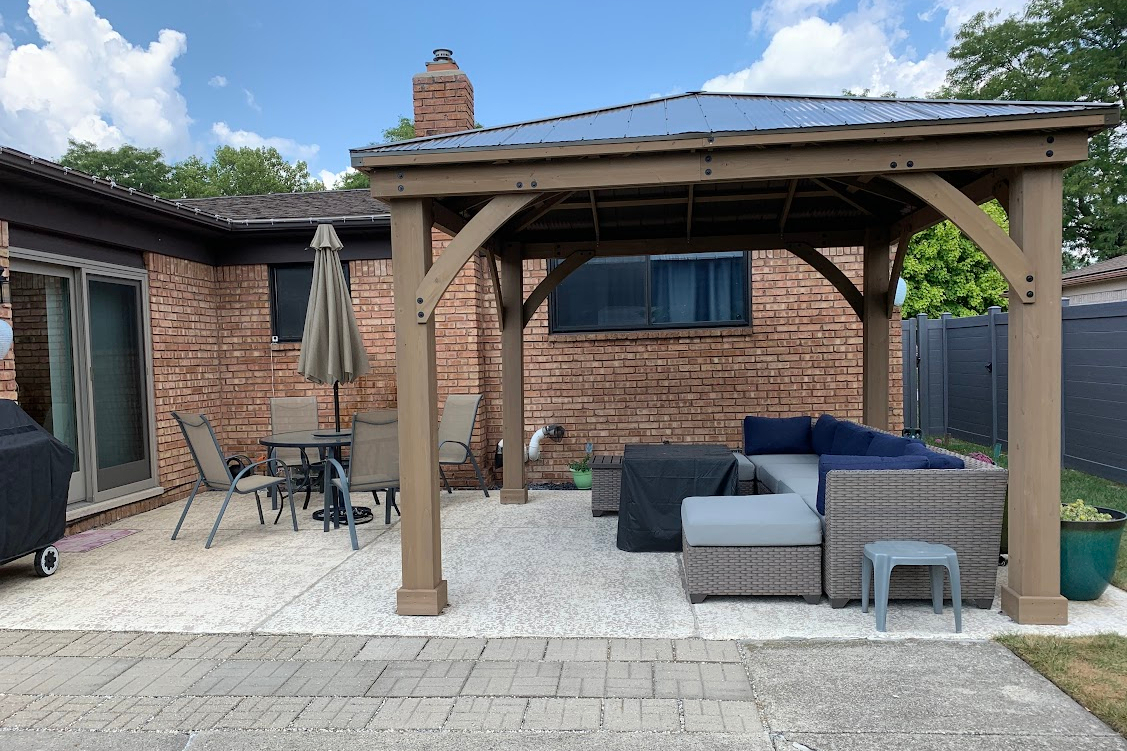Backyard patio under a wooden gazebo with a cushioned sectional, coffee table, and a dining set with umbrella against a brick wall.