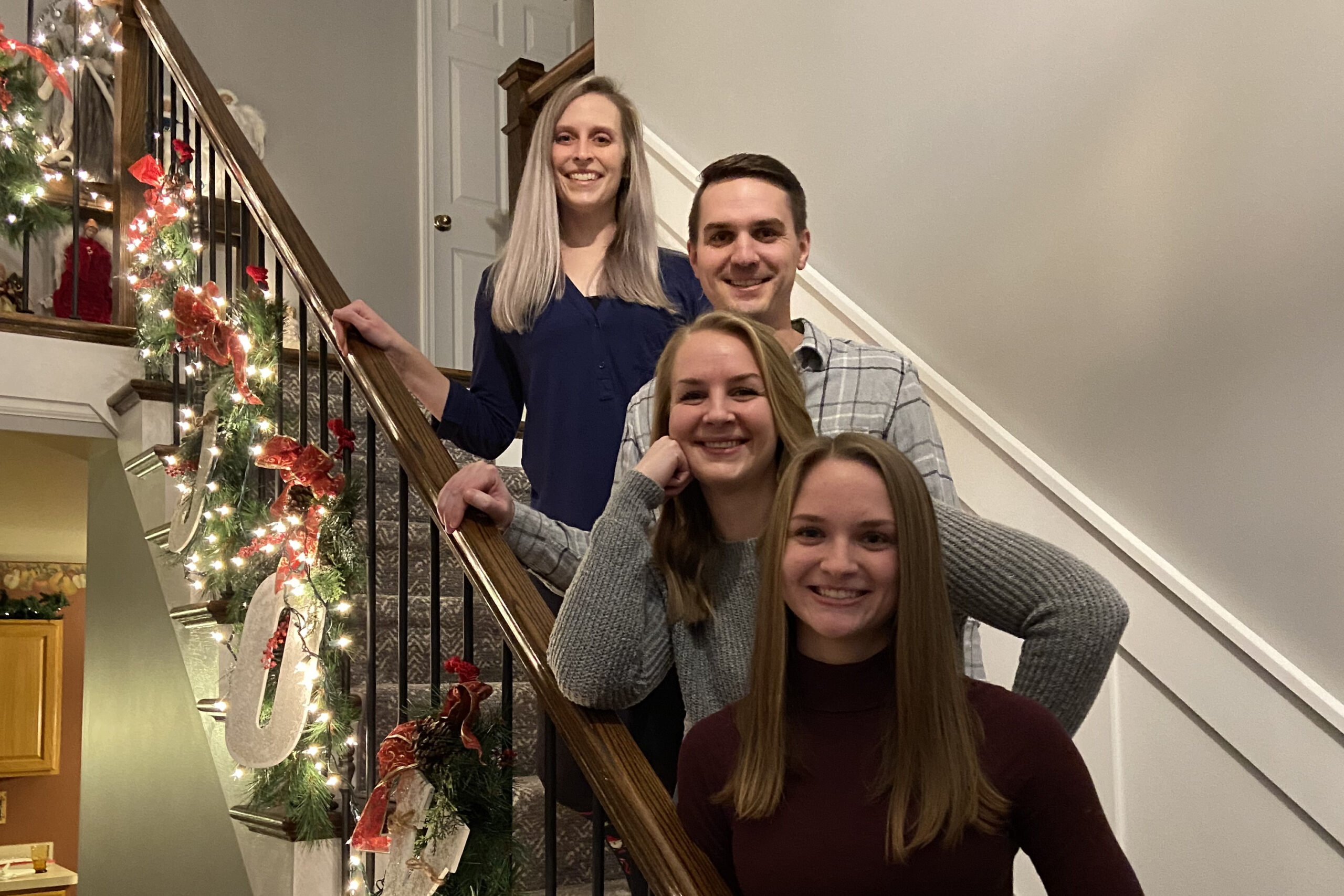 Group of four smiling people posing on a decorated staircase with Christmas lights and garland in the rail area.