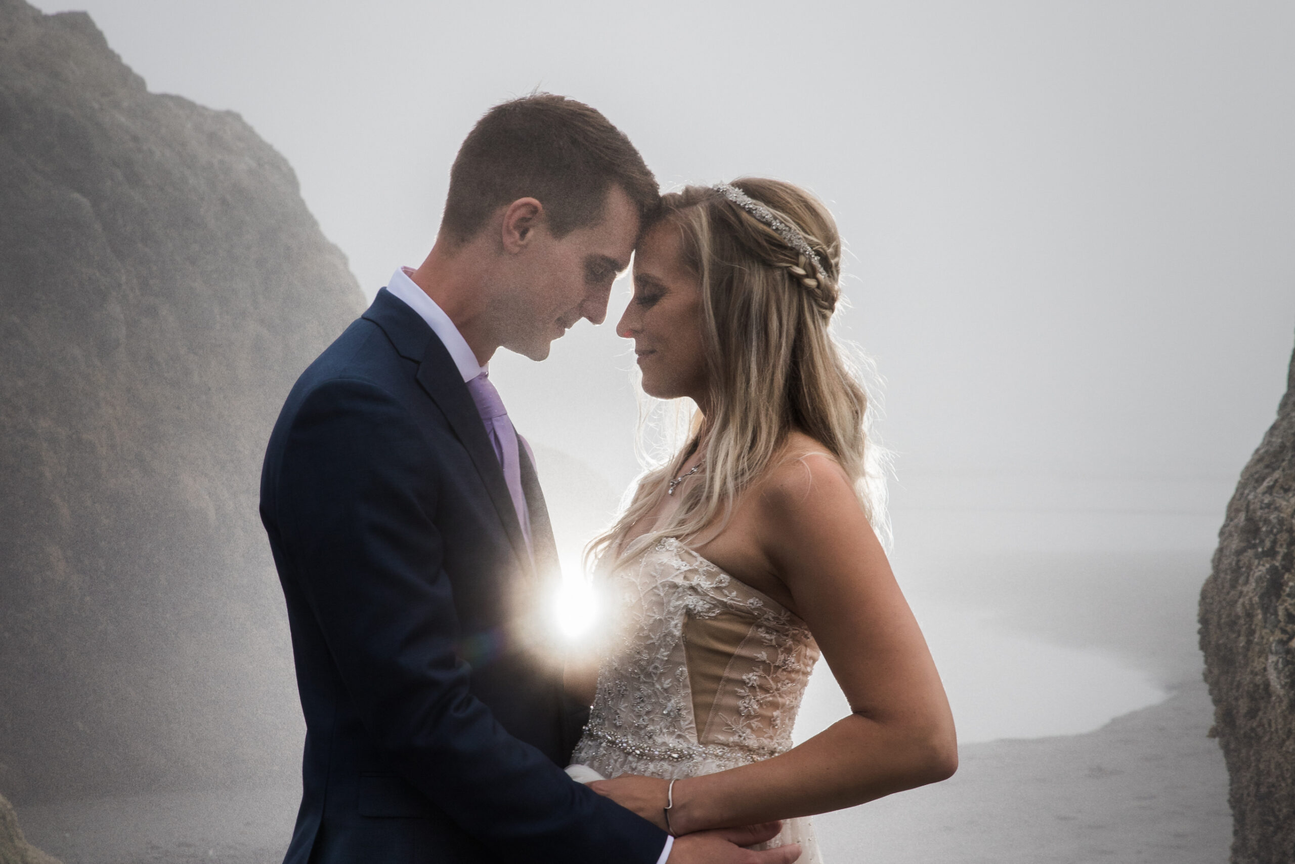 Bride in lace dress and groom in a dark suit embrace on a misty beach with rocky cliffs on either side of them at sunsetlight.