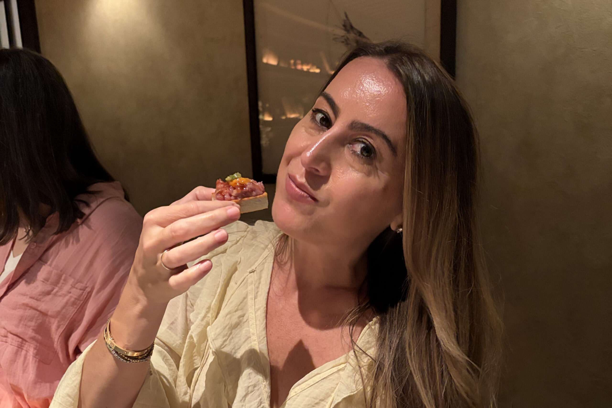 Smiling woman with long hair holds a small bite-sized appetizer near her lips in a warm, dim restaurant.