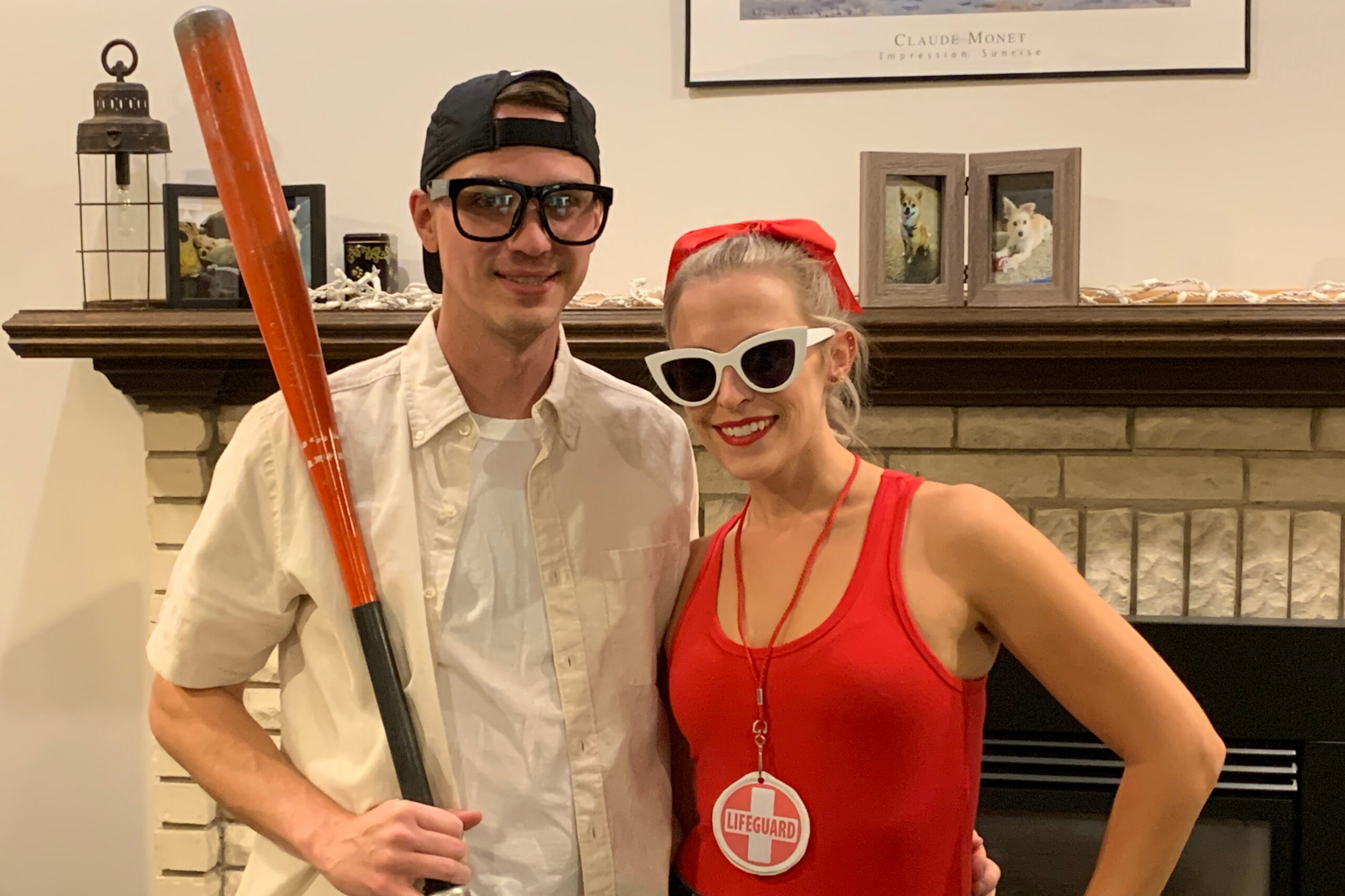 Two adults in costumes posing indoors by a fireplace, man with a long orange prop bat and backward cap, woman in a red lifeguard outfit with white sunglasses.