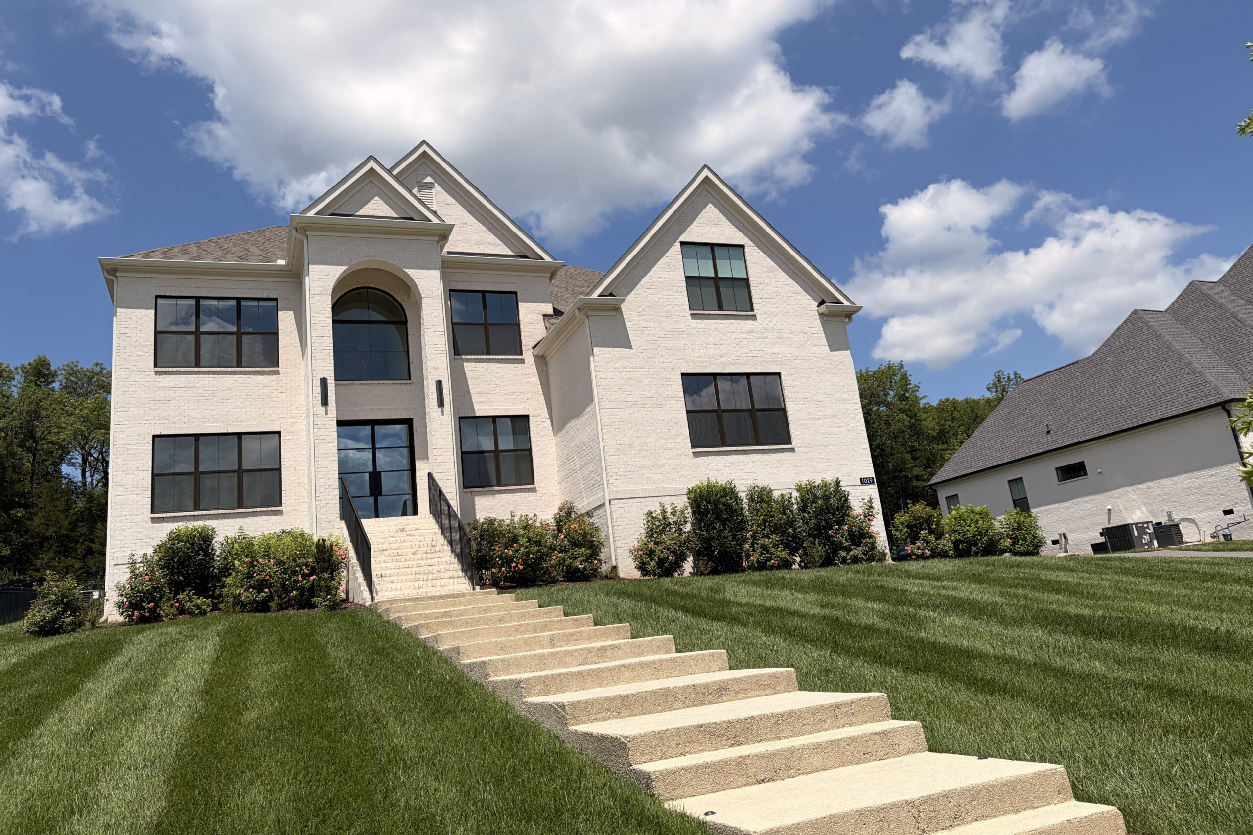 Modern white brick house with large black-framed windows and a curved entry arch on a manicured lawn under a blue sky with clouds.