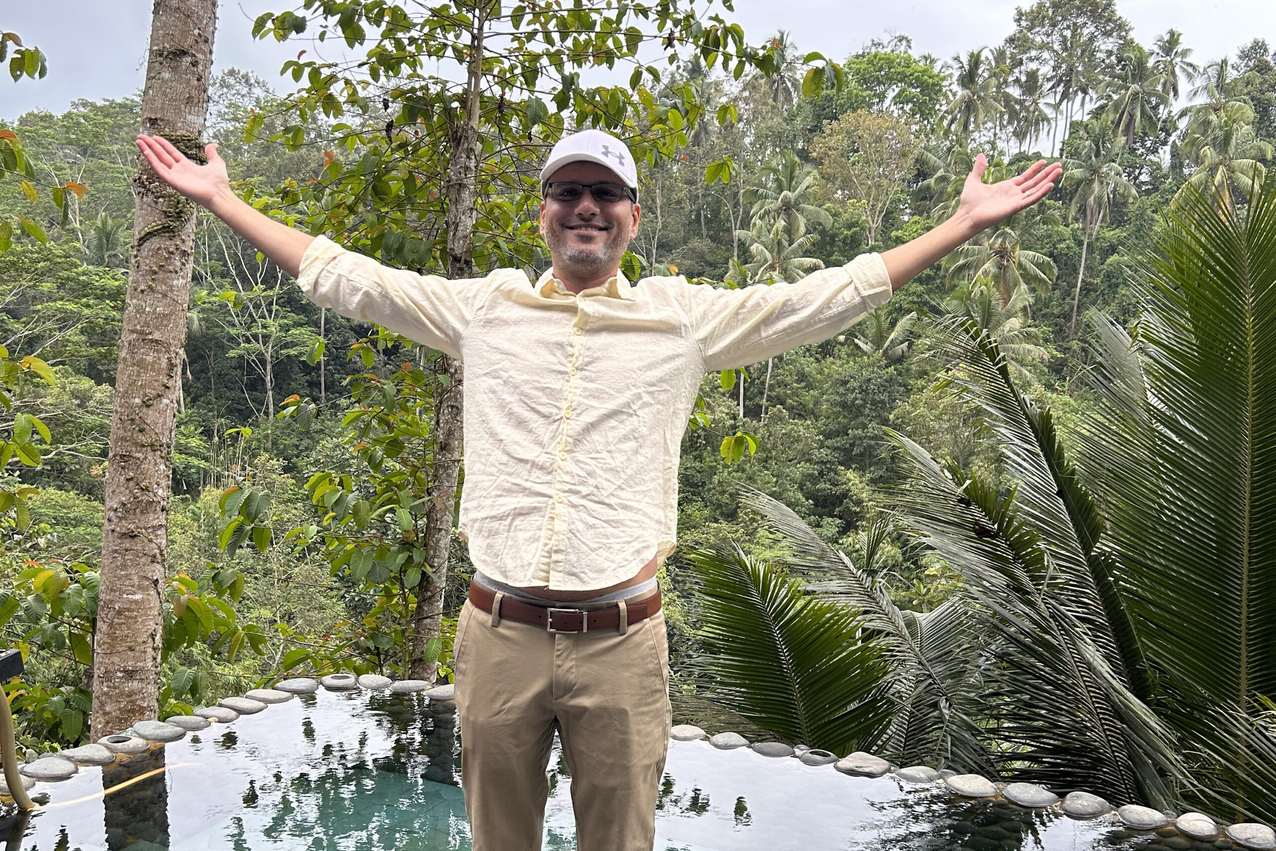 Man with arms raised in a tropical garden beside a reflecting pool, smiling at the camera.