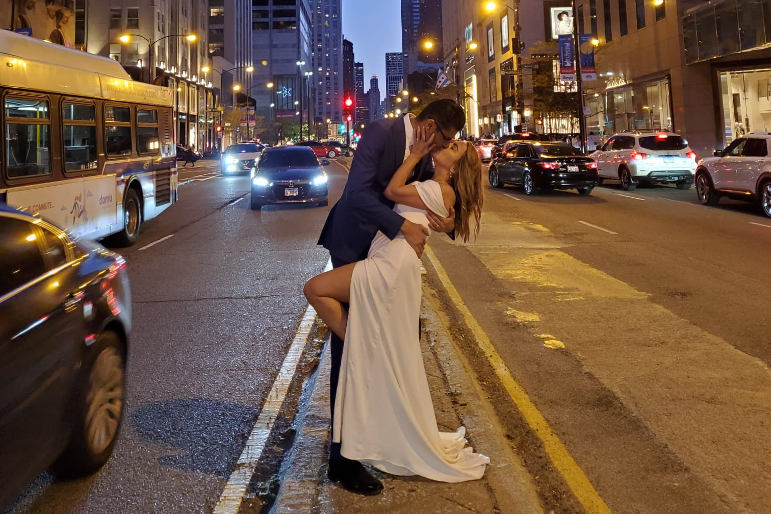 A man in a suit embraces a woman in a white wedding dress, kissing on a busy city street at night with traffic around them.