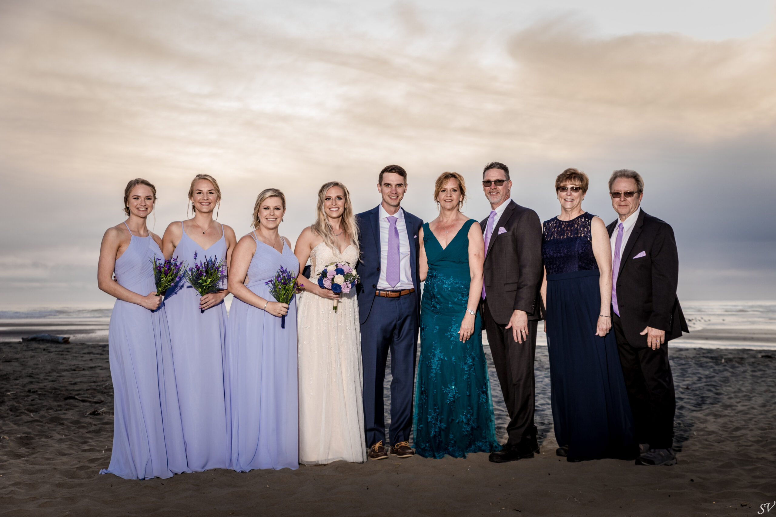 Group of wedding party on a beach at sunset: bridesmaids in lavender dresses, the bride in white, and guests in formal attire posing together.