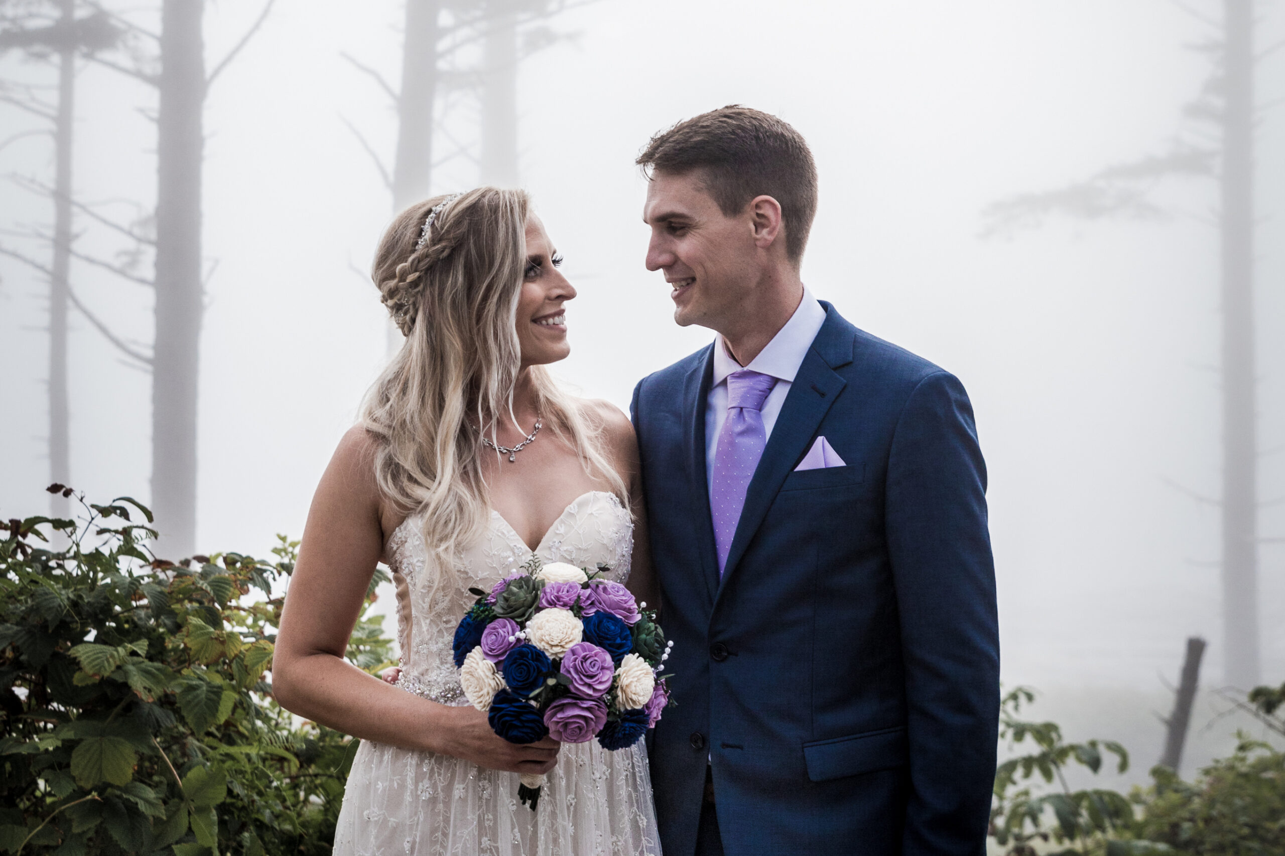 Bride and groom smile at each other in a foggy forest; bride in a white lace dress holding a purple-and-cream bouquet, groom in a navy suit.