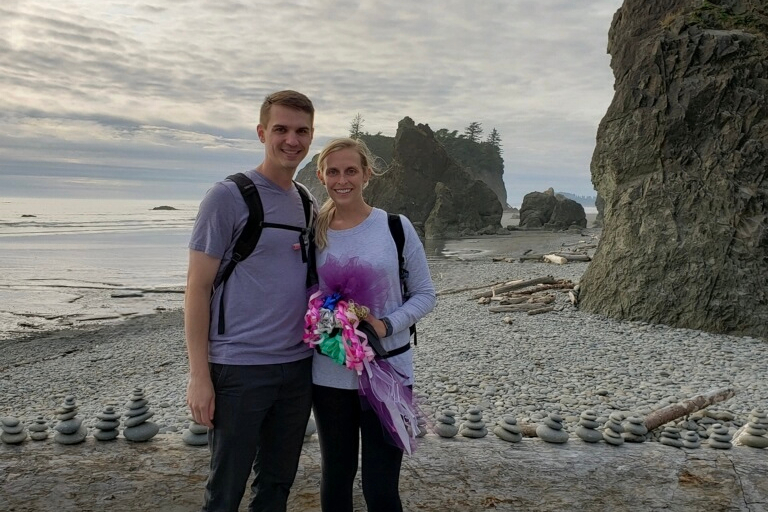 Smiling couple stands arm in arm on a pebble beach with stacked stones and rocky cliffs in the background, holding a colorful bouquet of flowers.