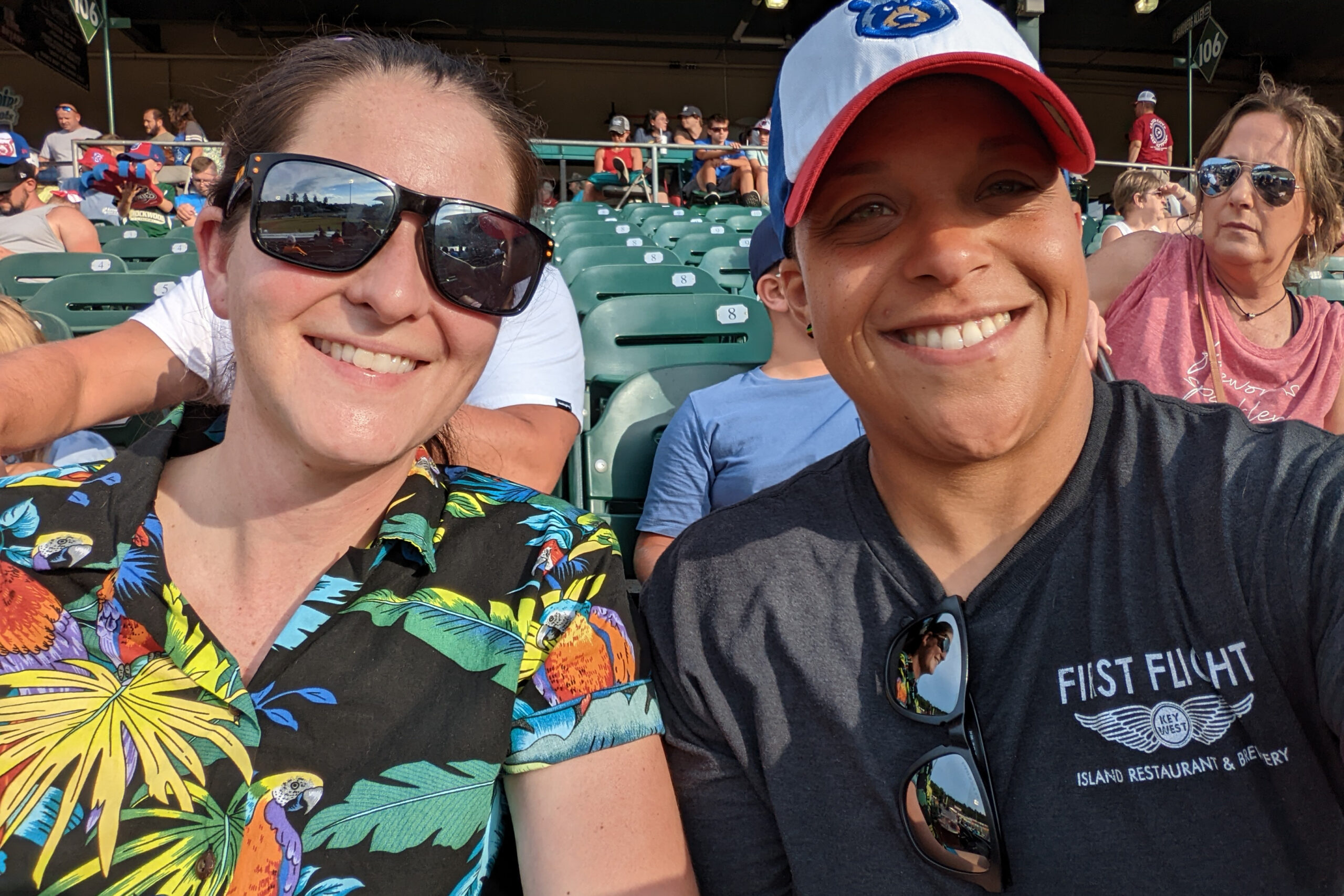 Two friends at a sports stadium taking a selfie; one in a colorful tropical shirt wearing sunglasses, the other in a dark T‑shirt with a cap and sunglasses on the shirt strap behind them.