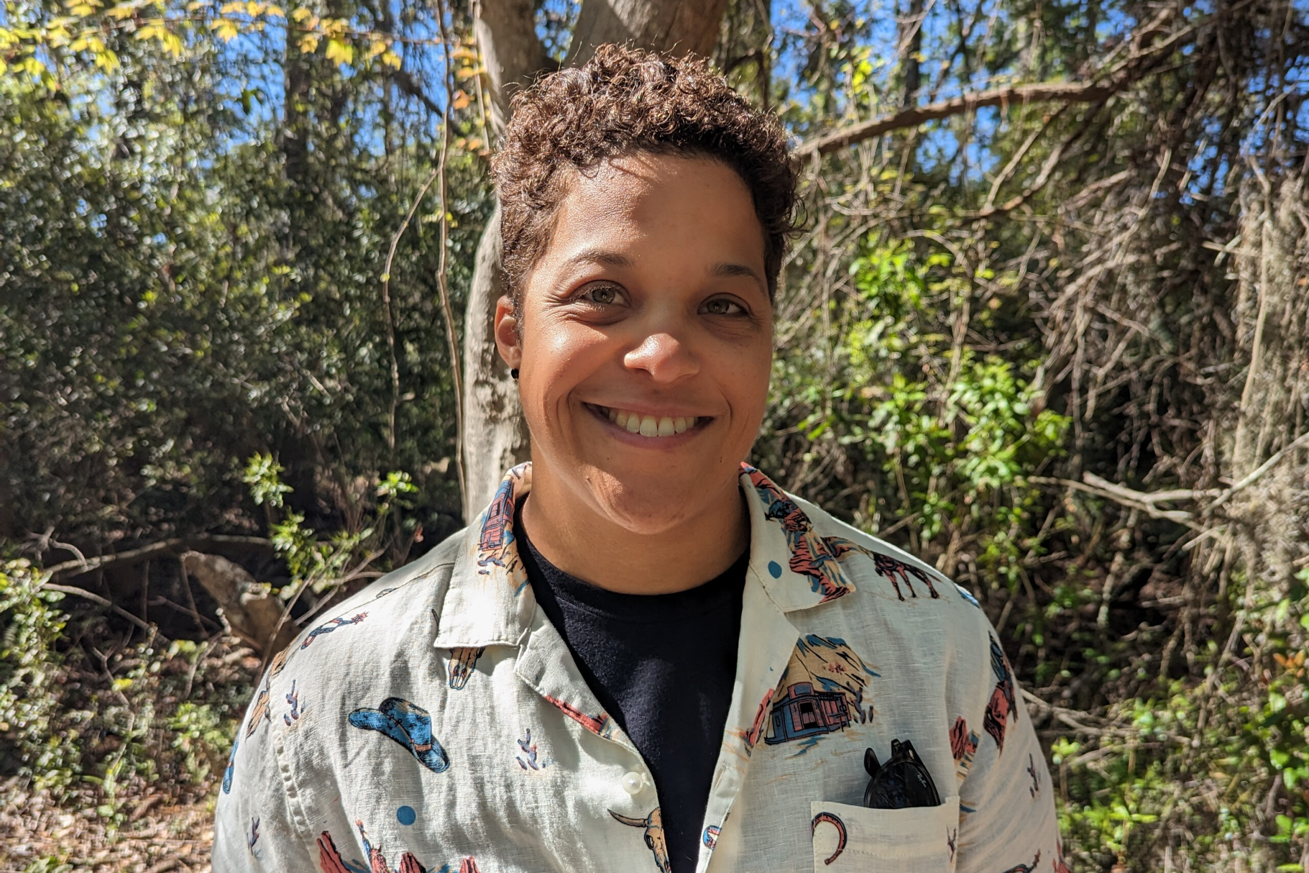 Close-up portrait of a smiling person outdoors in bright sunlight, wearing a light patterned shirt and sunglasses in the shirt pocket.