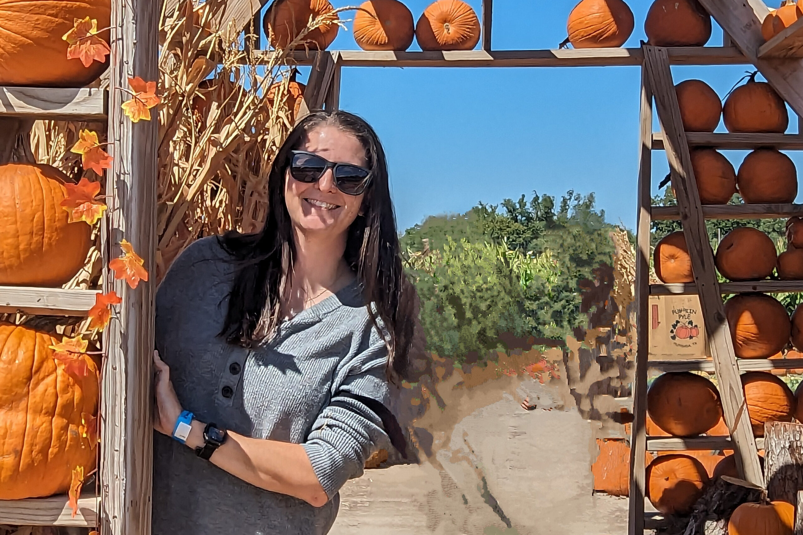 Smiling woman in sunglasses stands among stacked pumpkins on a wooden display at a fall pumpkin patch.
