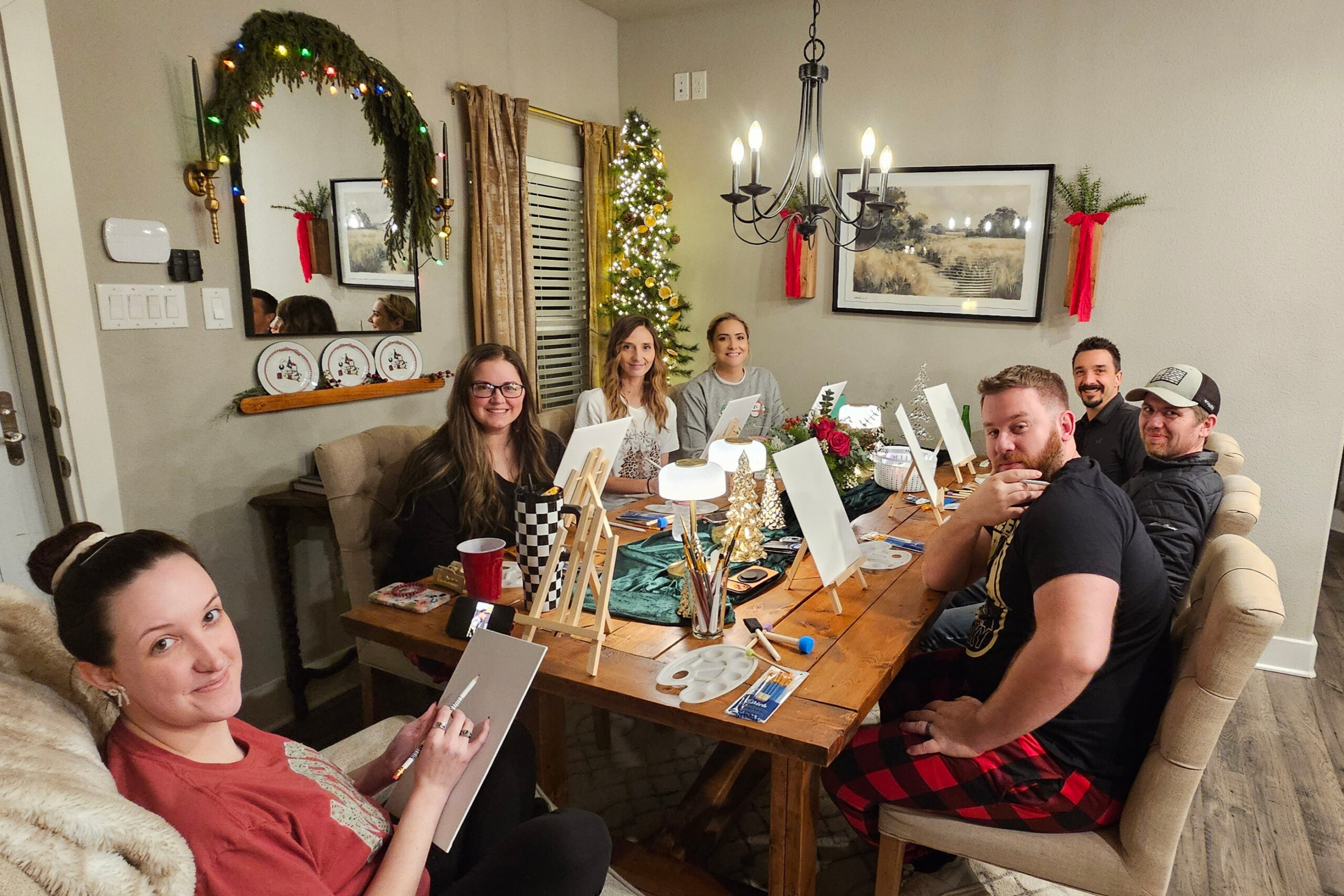 A group of friends gathered around a wooden dining table working on Christmas crafts, with easels, paints, and small canvases in a cozy, festively decorated living room.