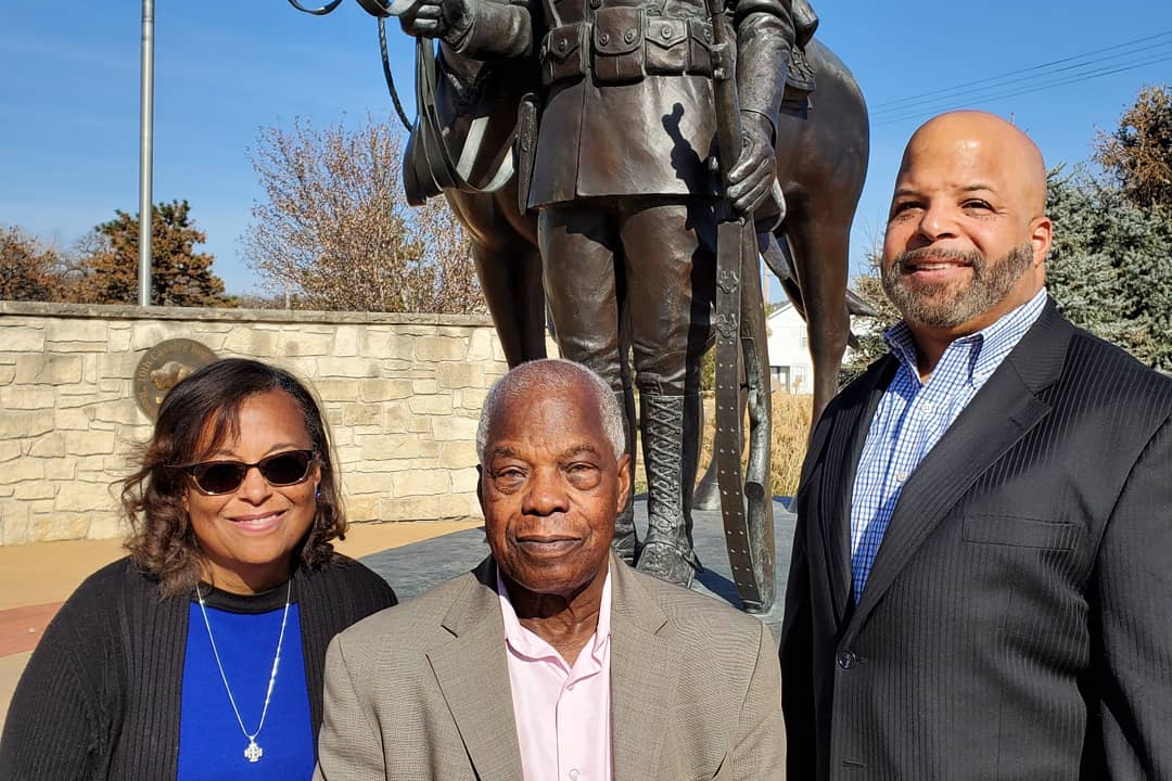Three adults pose smiling in front of a large equestrian statue outdoors on a sunny day.