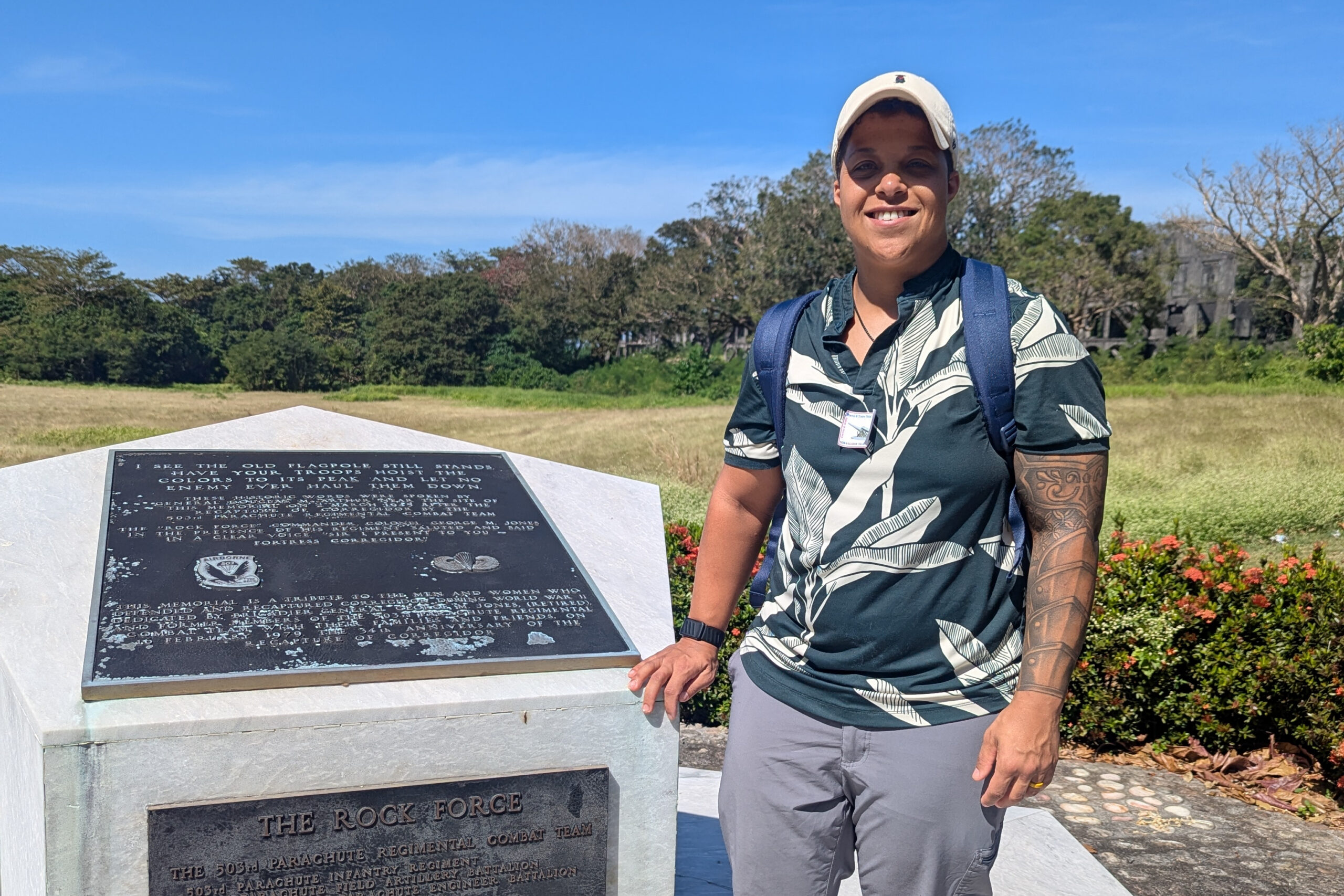 Smiling man with a backpack standing beside a granite memorial plaque on a sunny day in a park setting.