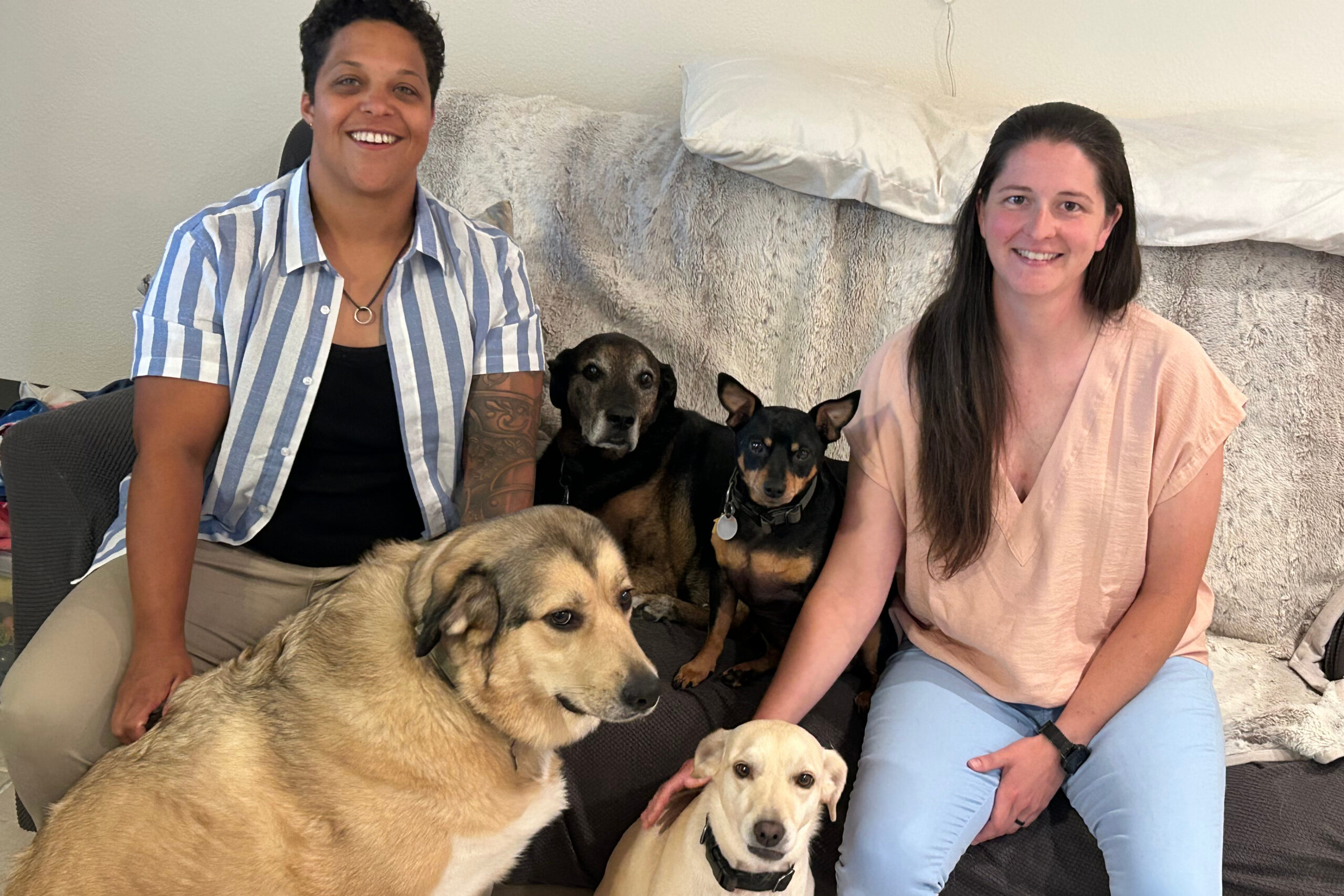 Two women sit on a couch posing with several dogs, smiling at the camera.