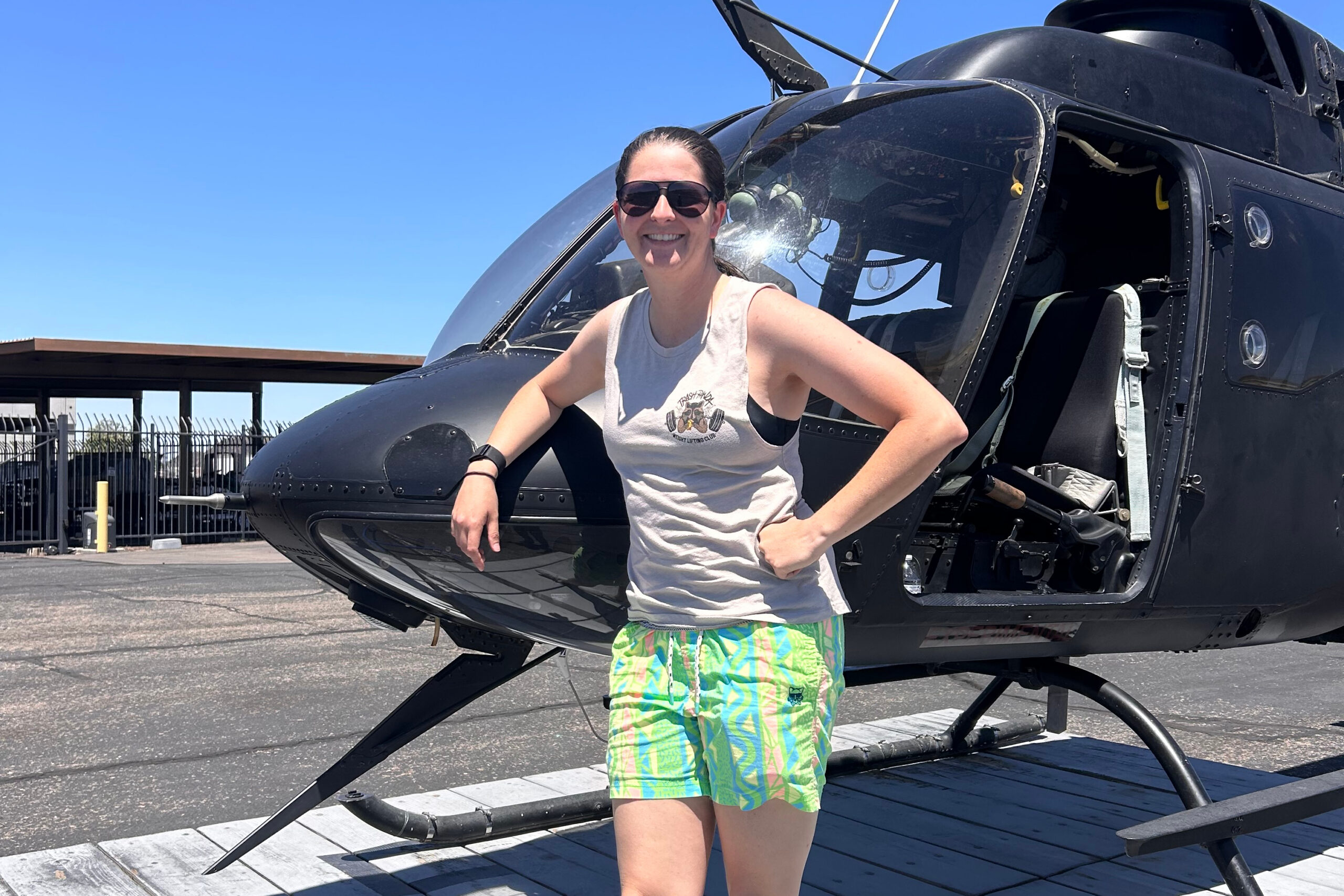 Smiling woman in sunglasses leans on a black helicopter with its cockpit open on a sunny day.