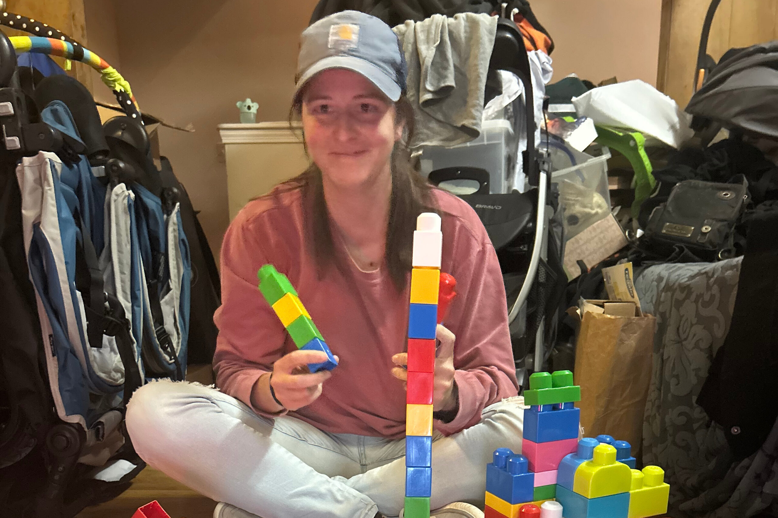 Person wearing a cap sits amid a cluttered room, building a tall colorful block tower with large plastic bricks.