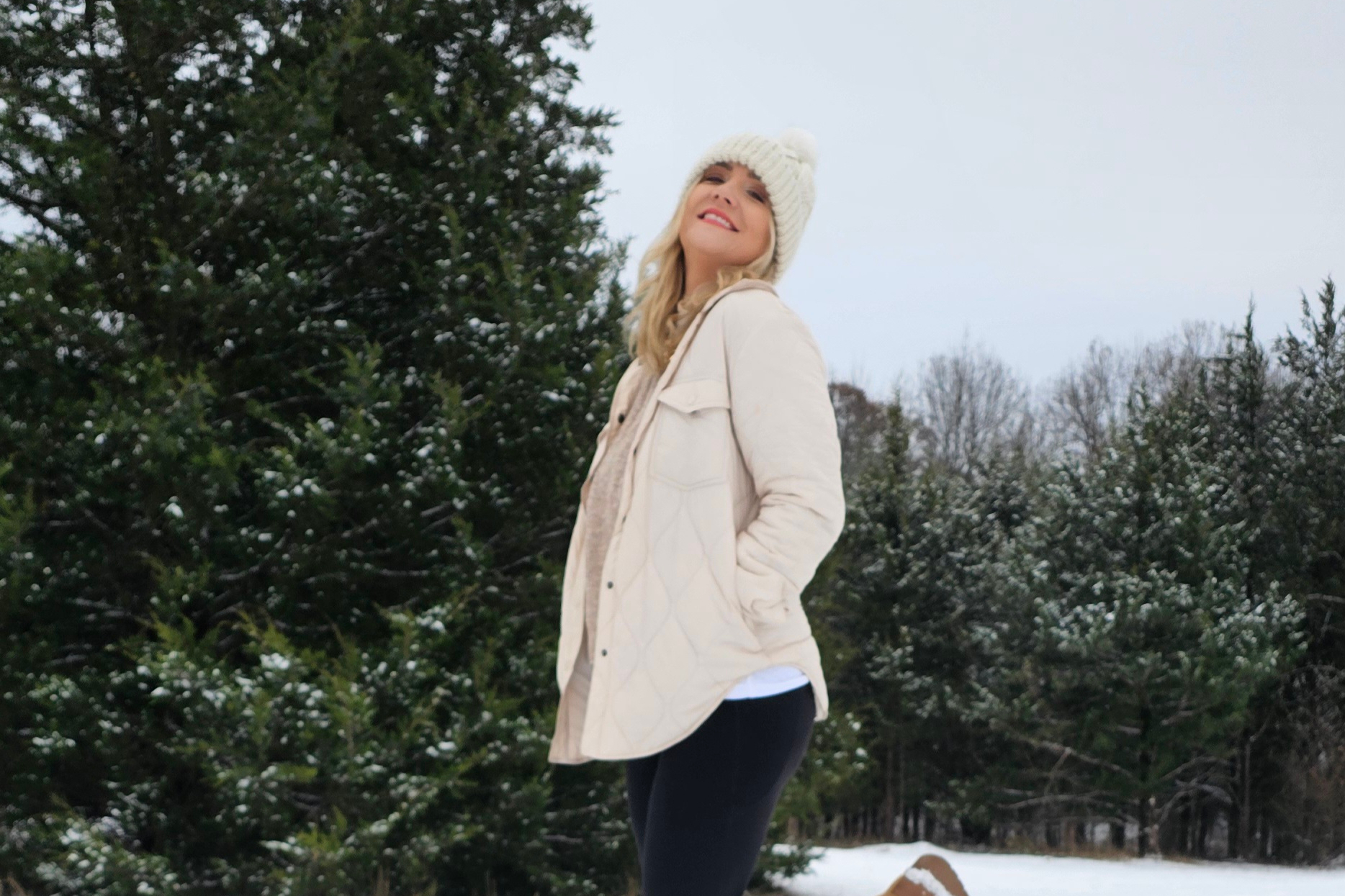 Woman in a beige quilted coat and white knit hat smiling outdoors in a snowy garden.