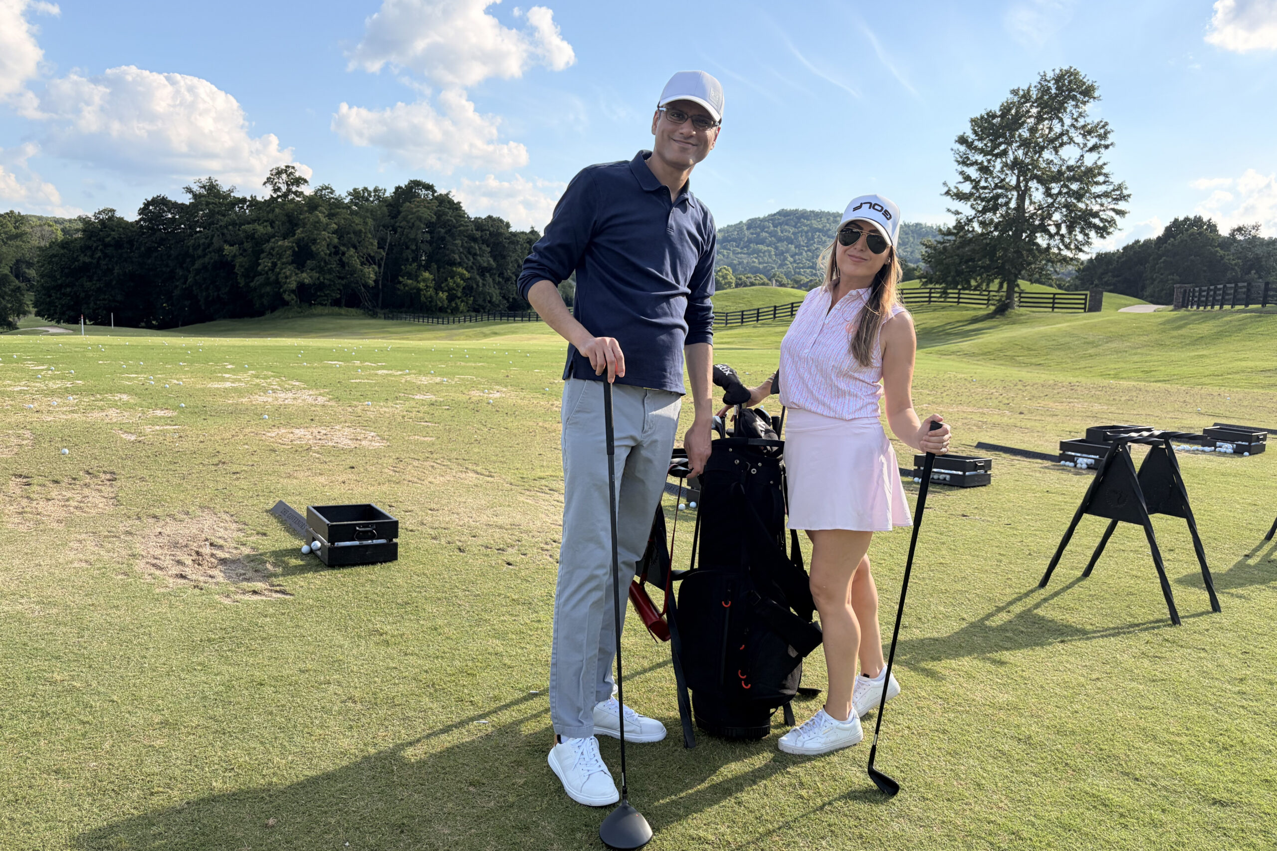 Two golfers posing with clubs on a sunny driving range, golf bags and scattered balls in front of them with trees in the background.