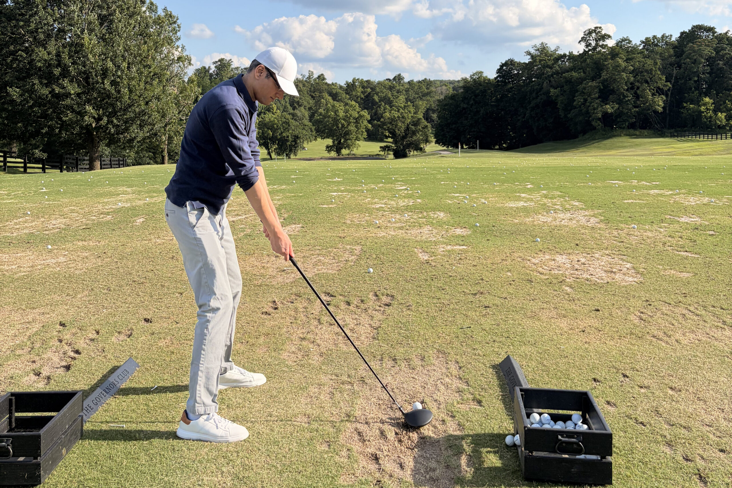 A man wearing a white cap and navy sweater tees off on a sunny driving range, with ball baskets nearby.