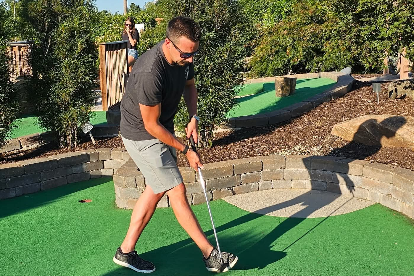 Man in dark t-shirt and gray shorts lining up a putt on a mini-golf green, with a woman watching in the background.