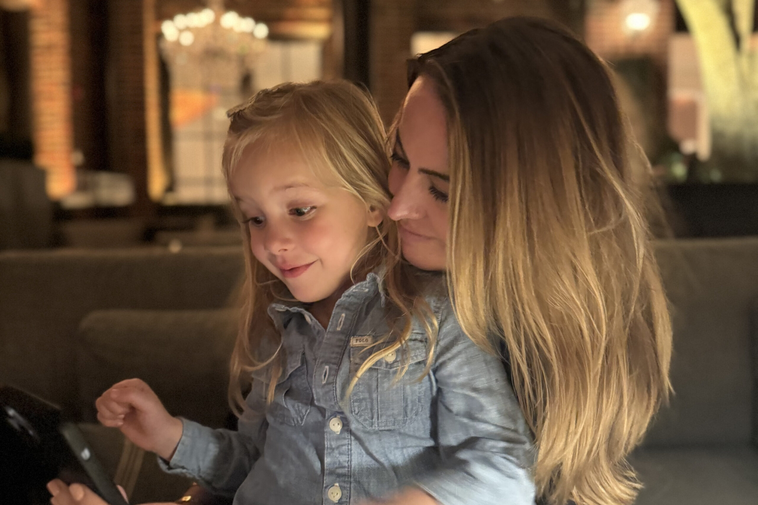 Woman with long hair and a young girl closely look at a tablet together in a warmly lit indoor setting.