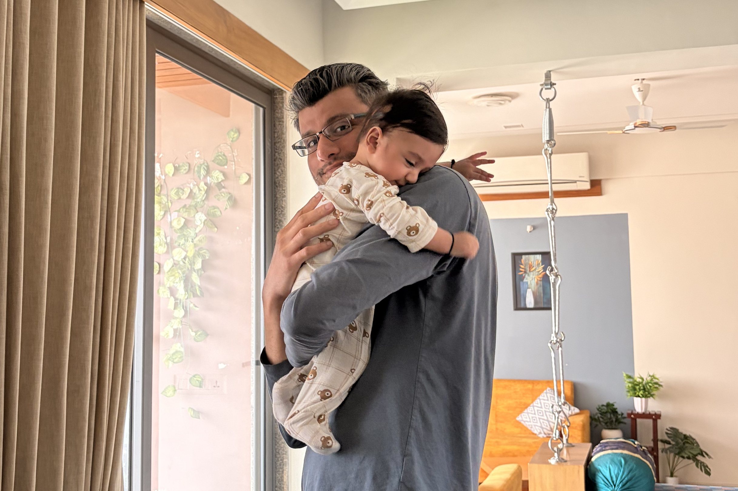 Father in glasses hugs a smiling baby indoors, near a window with beige curtains and plants in a cozy living room.