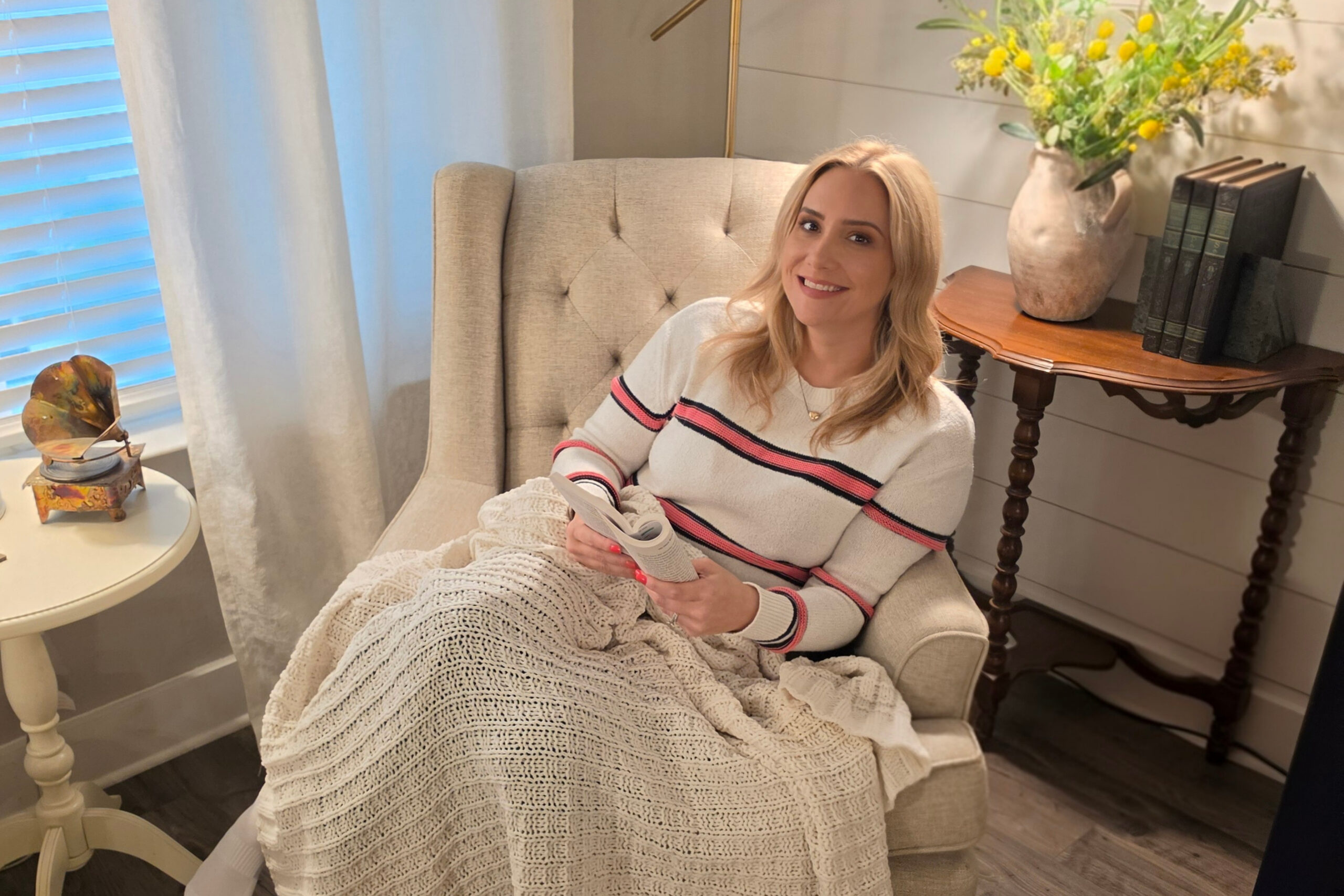 Woman relaxing in a tufted beige chair, knitting a cream blanket beside a side table with a vase of yellow flowers and books.