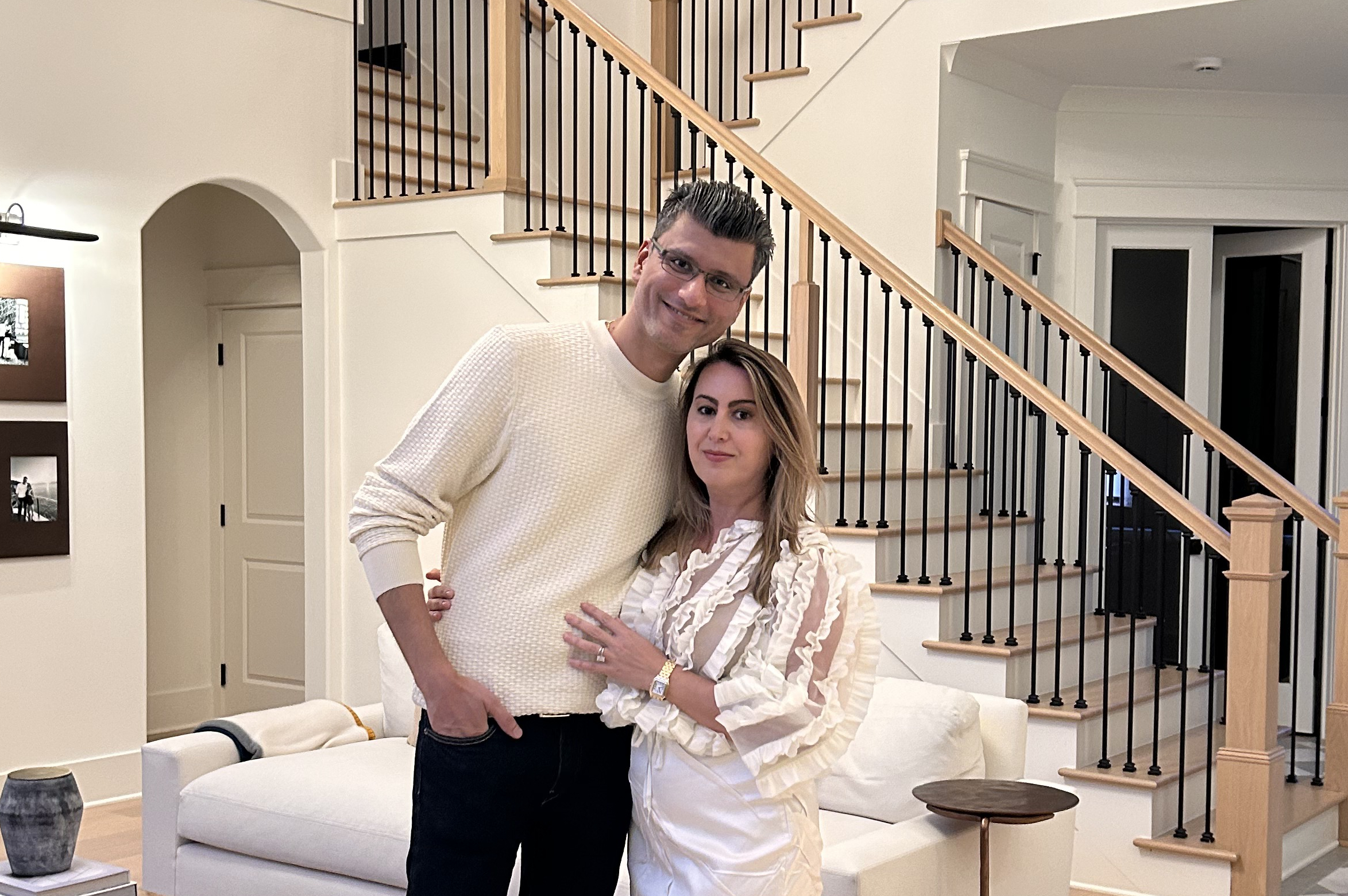 Man and woman posing together in a bright living room with a wooden staircase behind them.