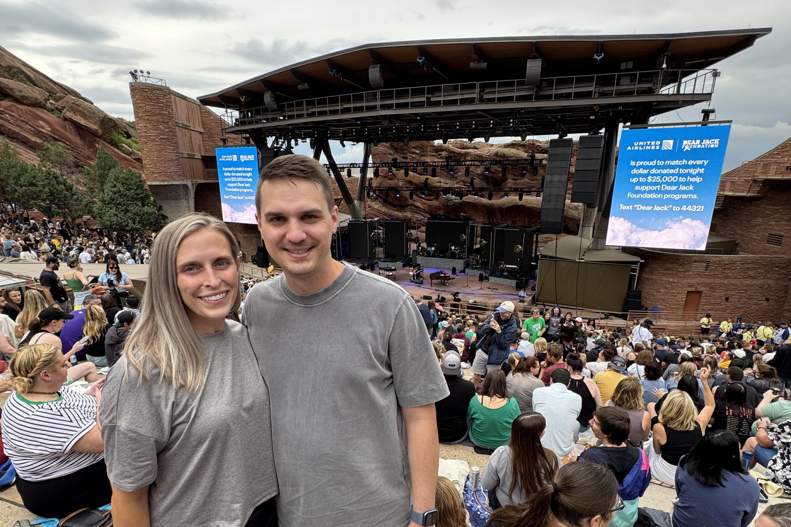 Smiling couple in gray shirts posing at a large outdoor concert venue with a stage and crowd behind them.