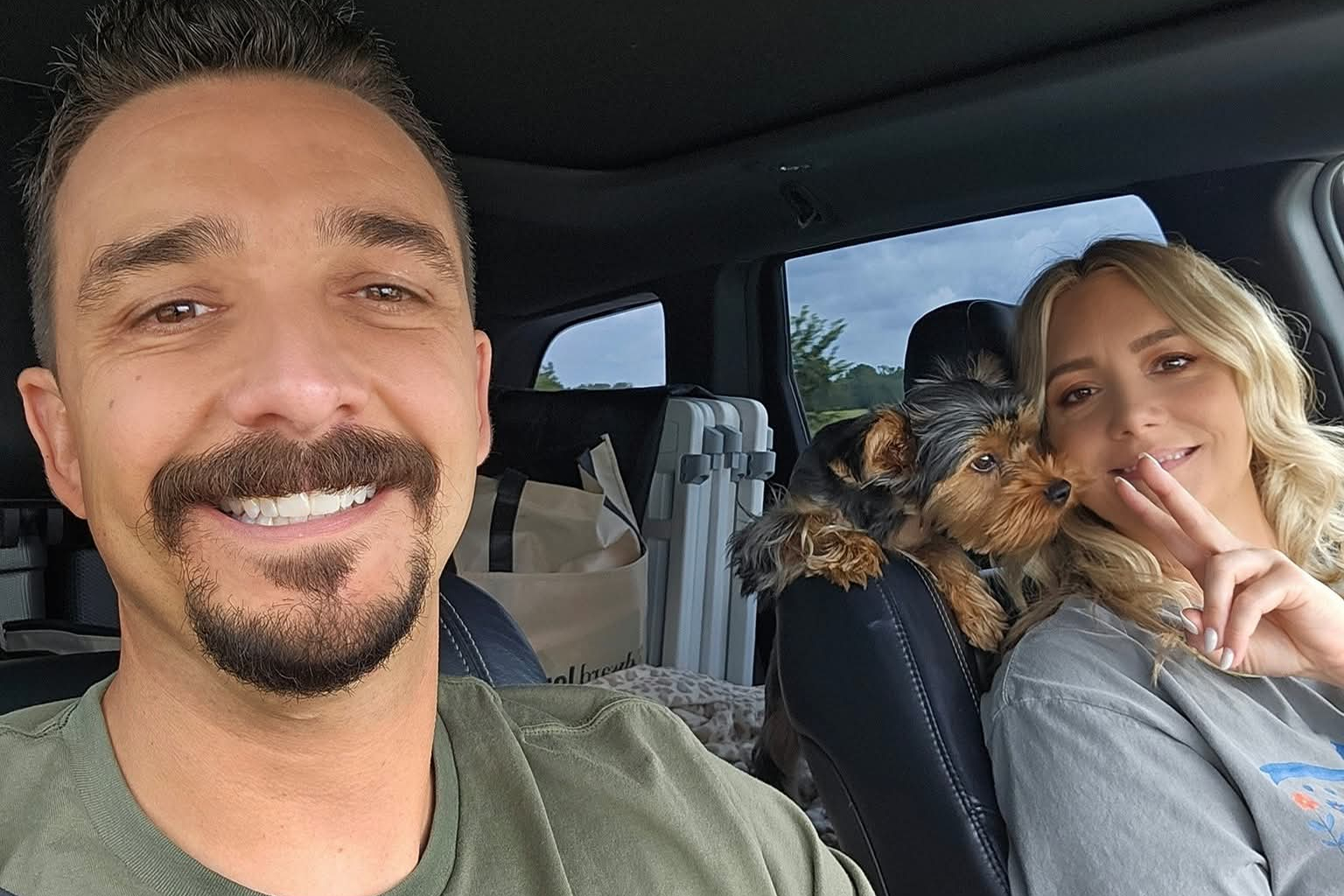 Smiling man in a car passenger seat with a small Yorkshire terrier on the woman’s shoulder in the driver’s seat; travel bags visible in back seat.