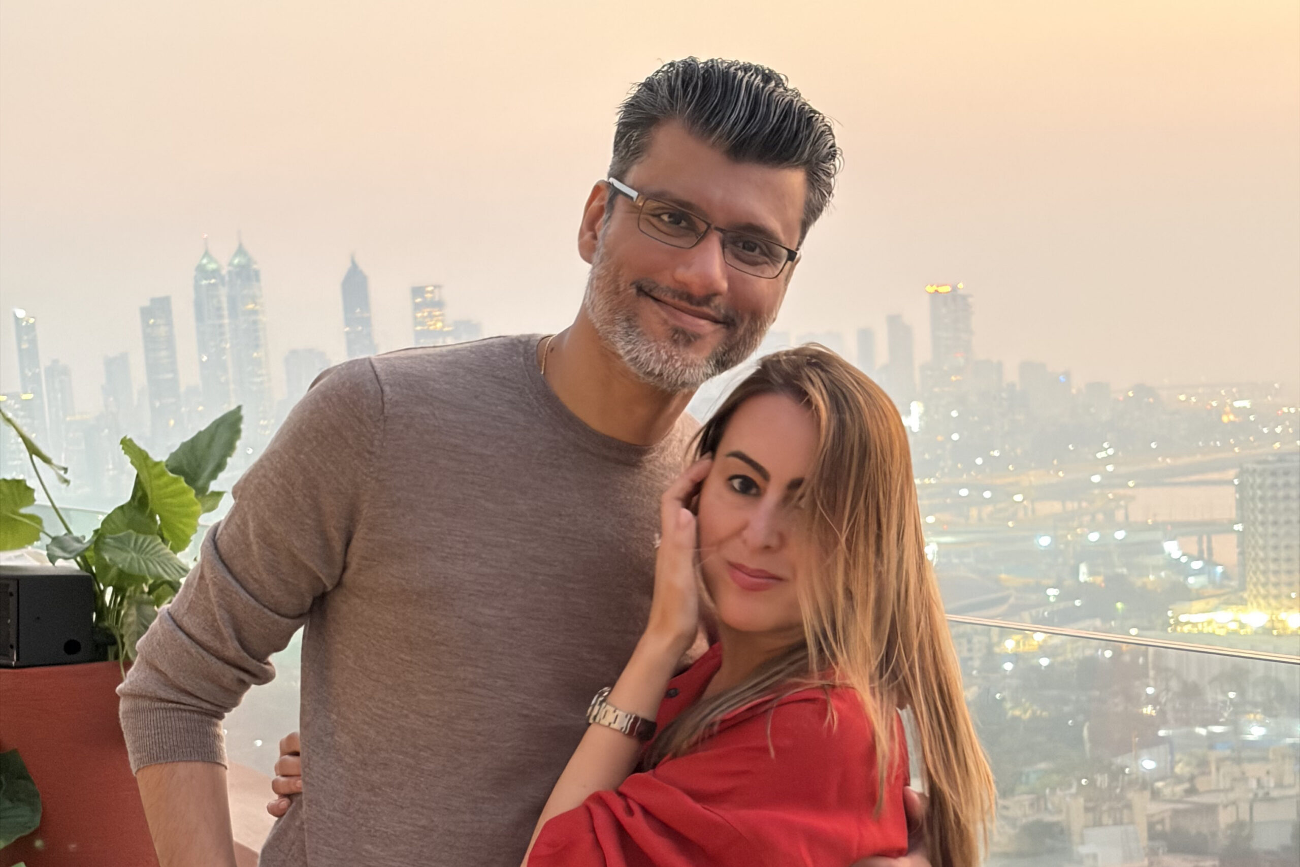 Smiling couple posing together on a balcony with a city skyline at sunset behind them