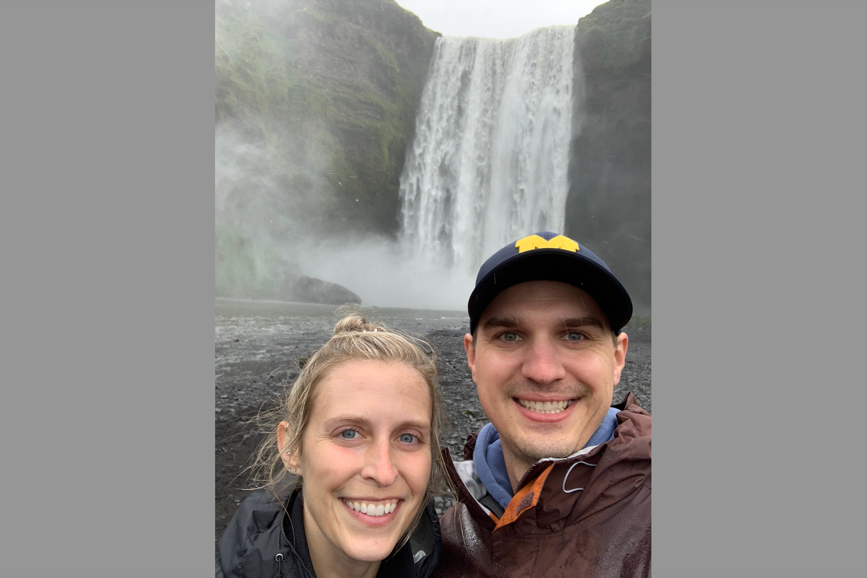 Smiling couple taking a selfie in front of a tall waterfall with mist rising from the pool below. Both wear outdoor jackets; woman in the foreground, man wearing a navy cap.