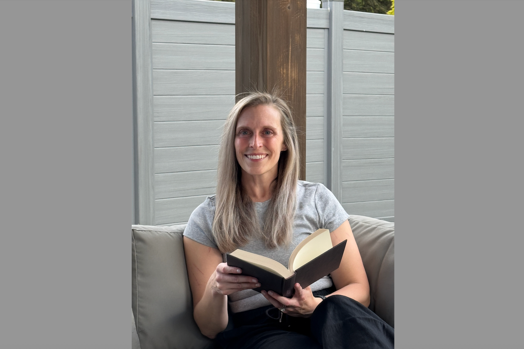 Woman with blonde hair sits on a patio couch, reading an open paperback book and smiling.
